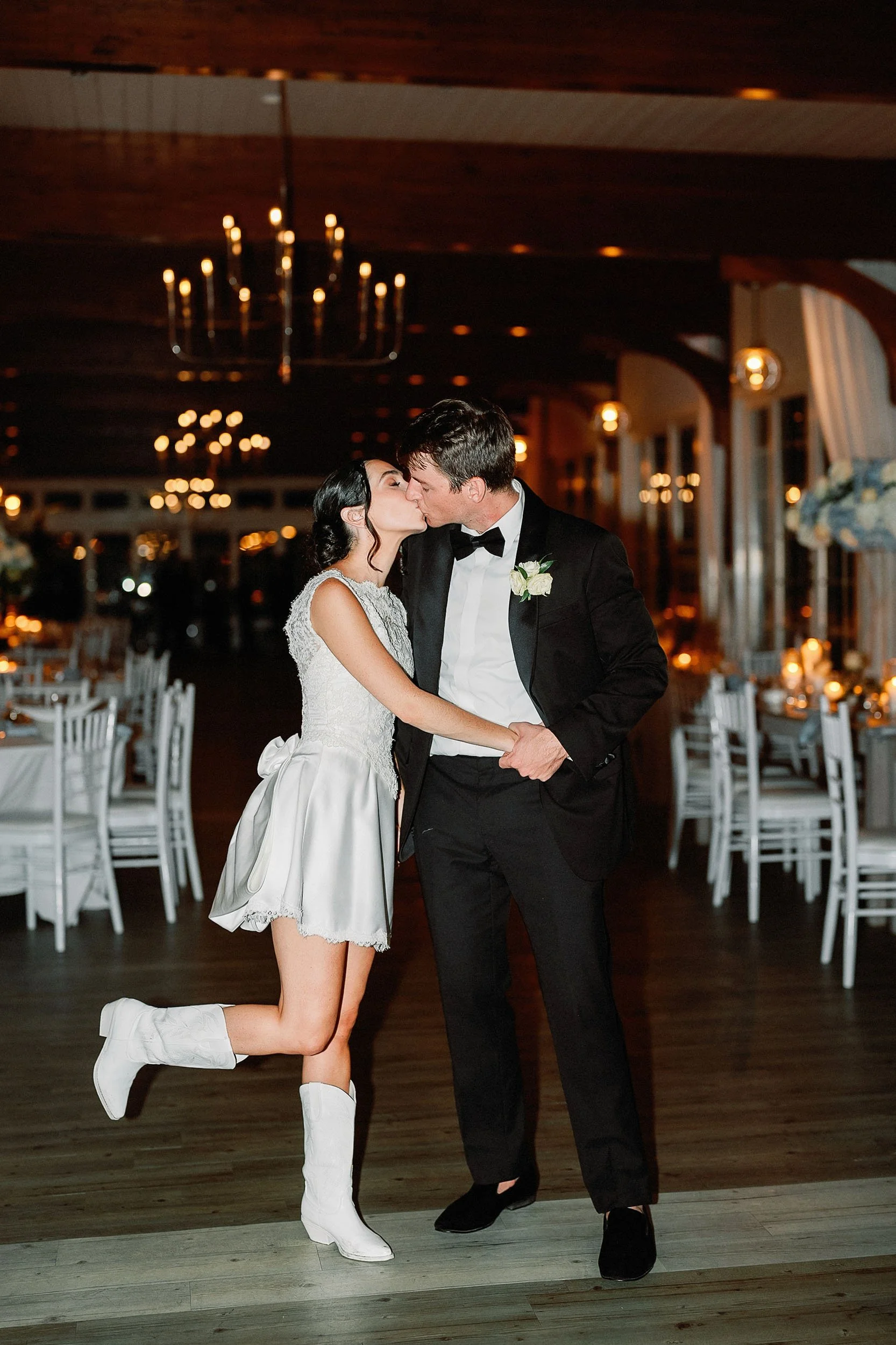 A couple dressed in formal wedding attire sharing a kiss at their wedding reception, with decorated tables and warm lighting in the background.