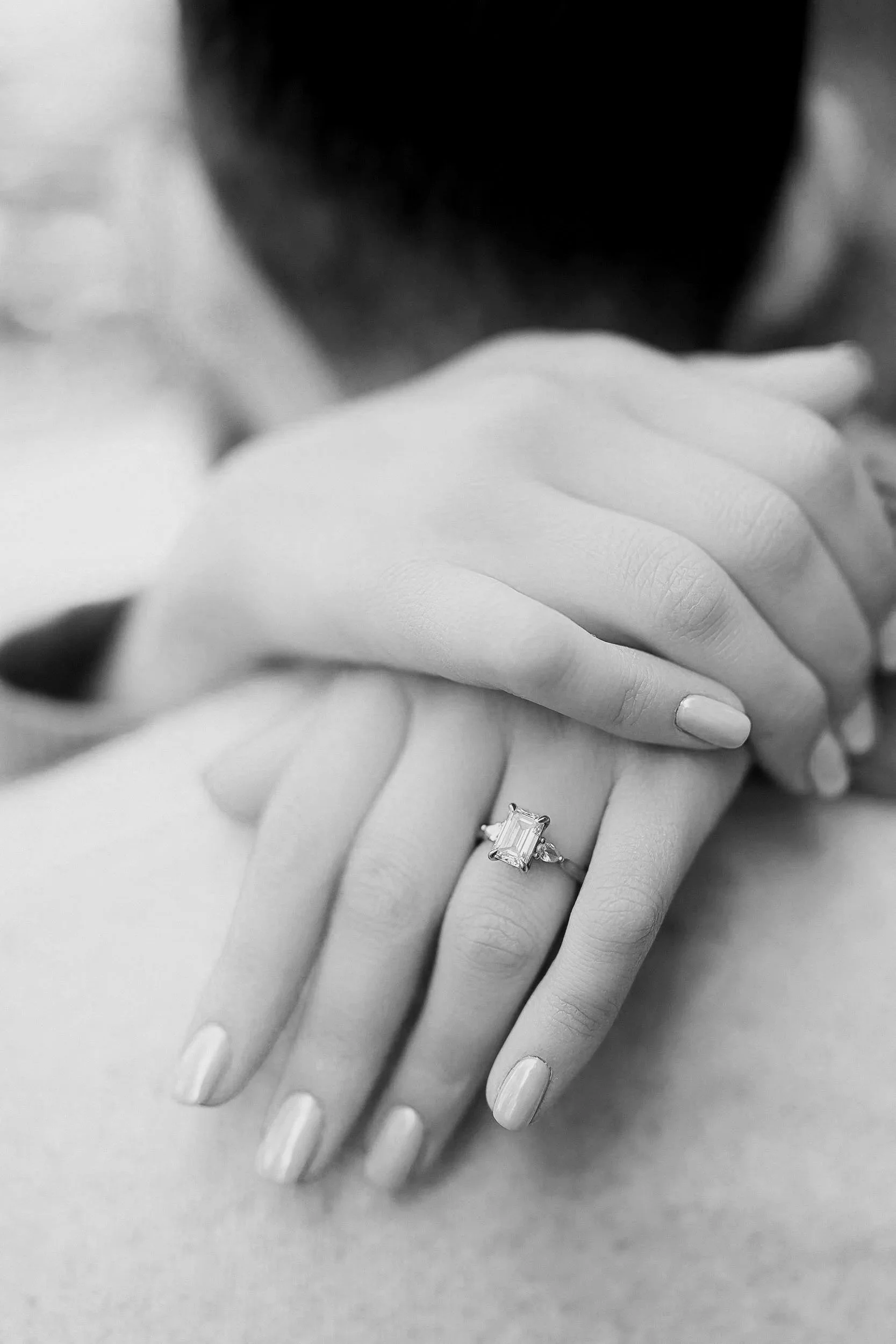 A woman with well-manicured nails shows her hand with an elegant rectangular-cut engagement ring with a halo setting.