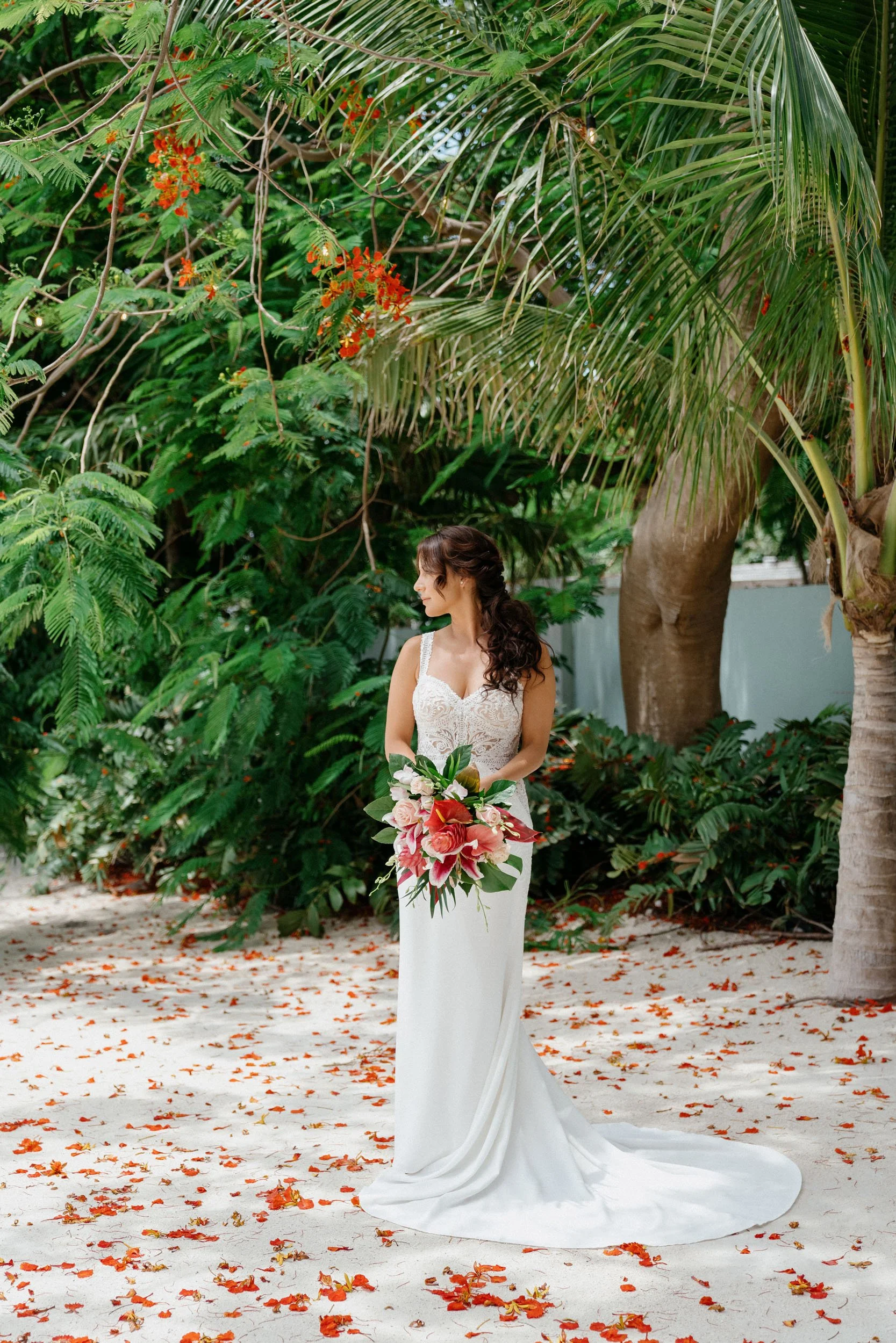 A woman in a white wedding dress holding a bouquet of pink and red flowers standing on a sandy path with scattered red leaves, surrounded by green tropical plants and palm trees.