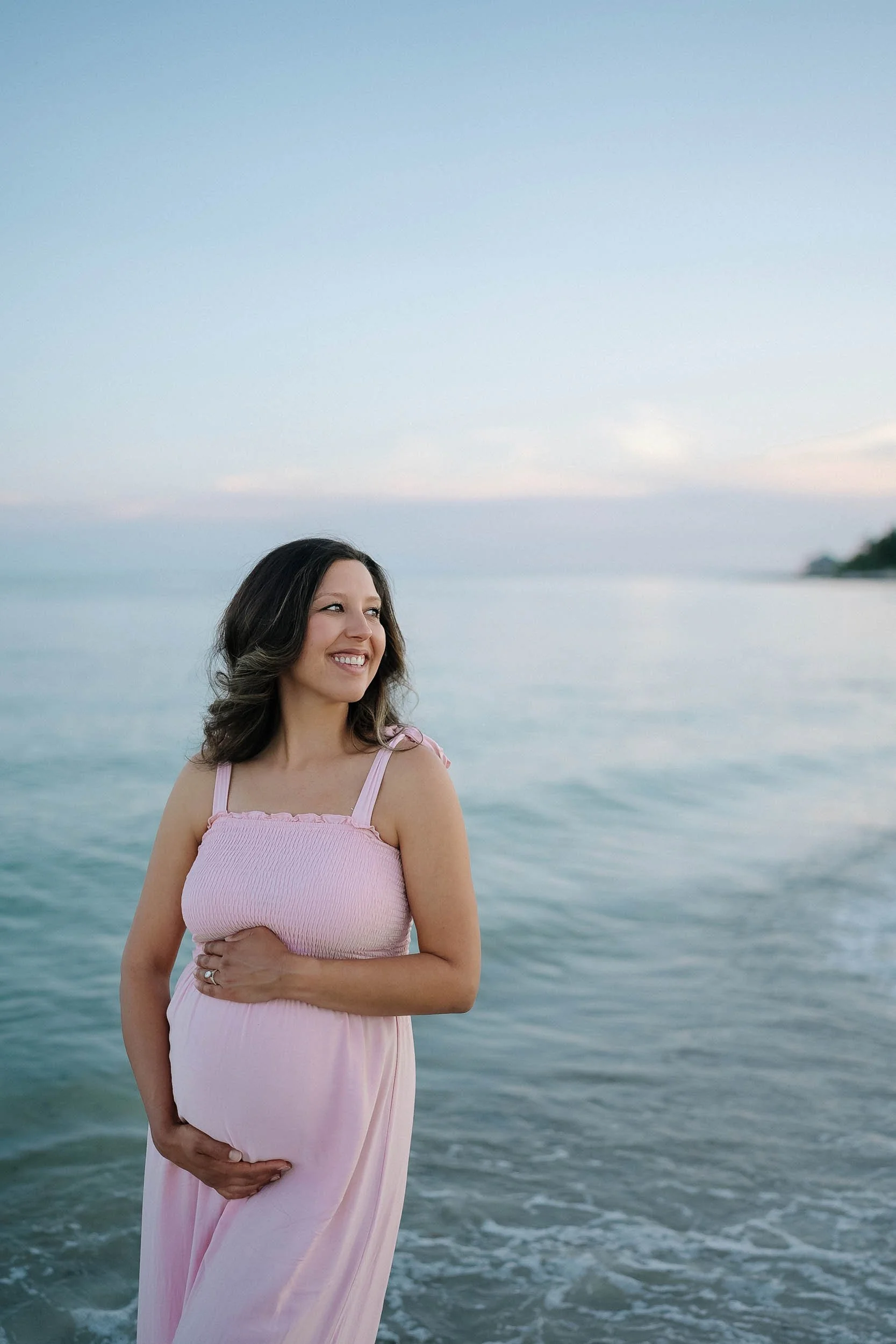 Pregnant woman in a pink dress standing in the water at the beach, smiling, with an ocean and sky in the background.