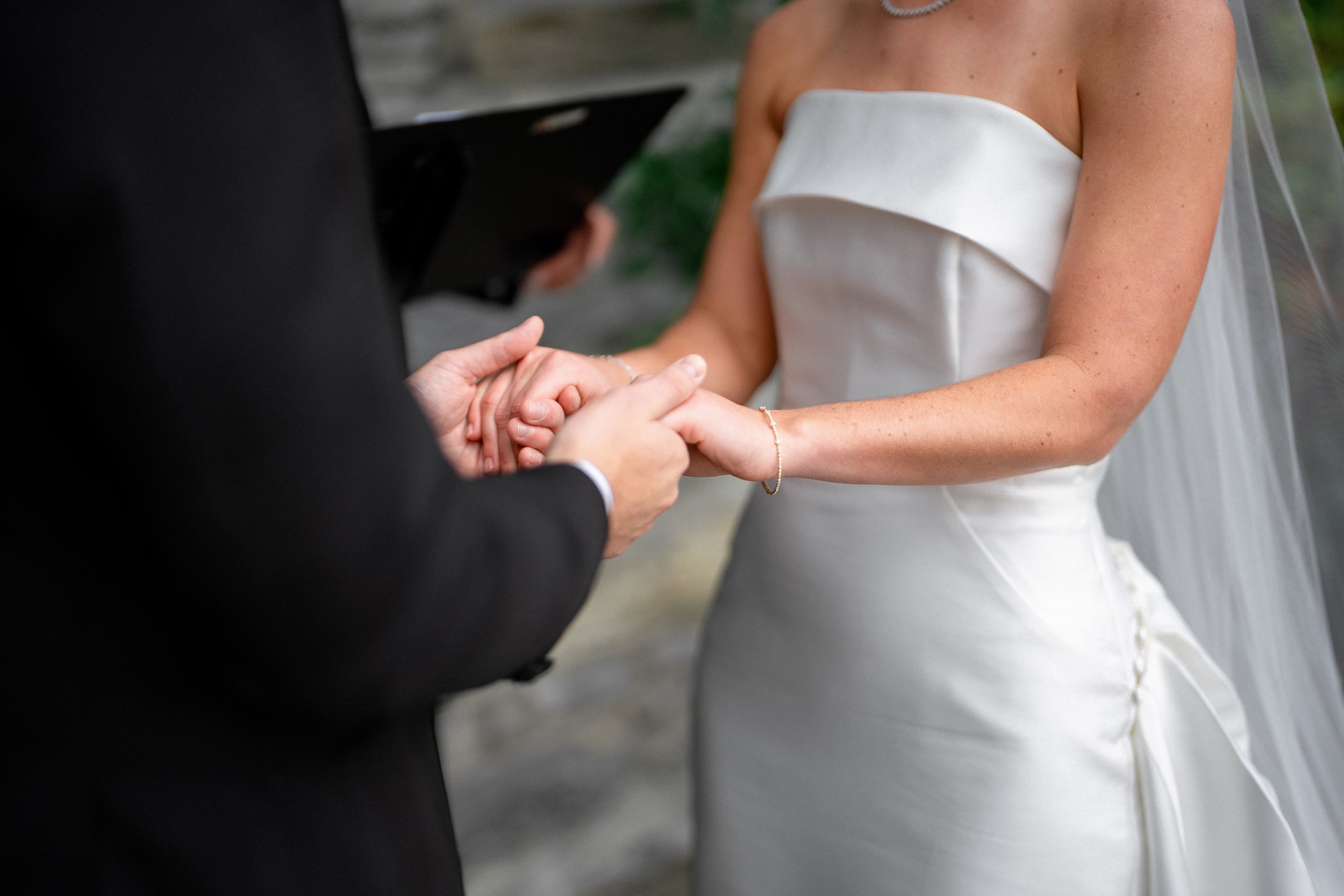 A bride and groom holding hands during a wedding ceremony, with the groom wearing a black suit and the bride in a white strapless wedding dress.