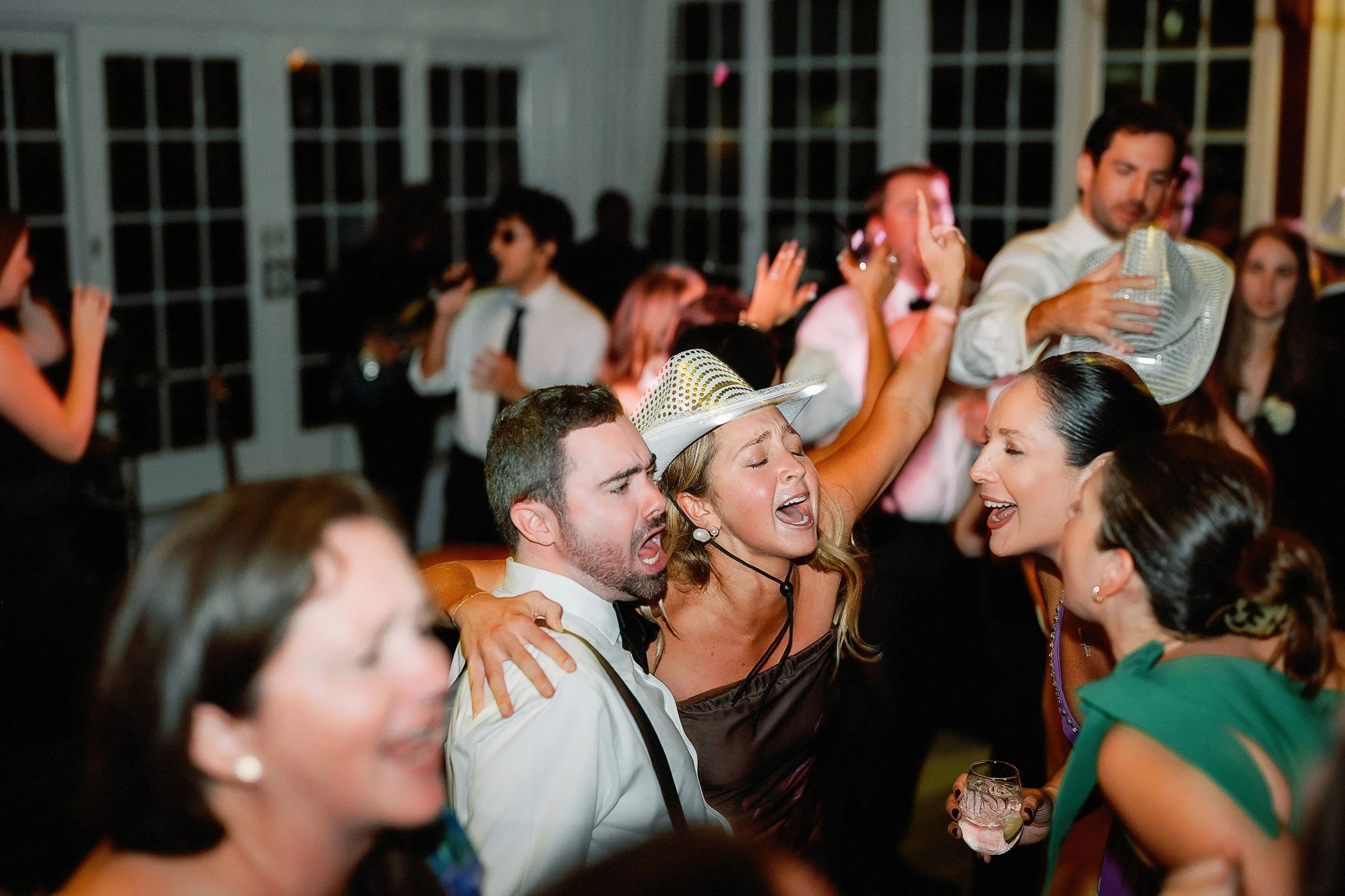 People at a lively party or celebration, singing or dancing with expressions of enjoyment, some holding drinks, and one woman wearing a hat, in an indoor venue with large windows.