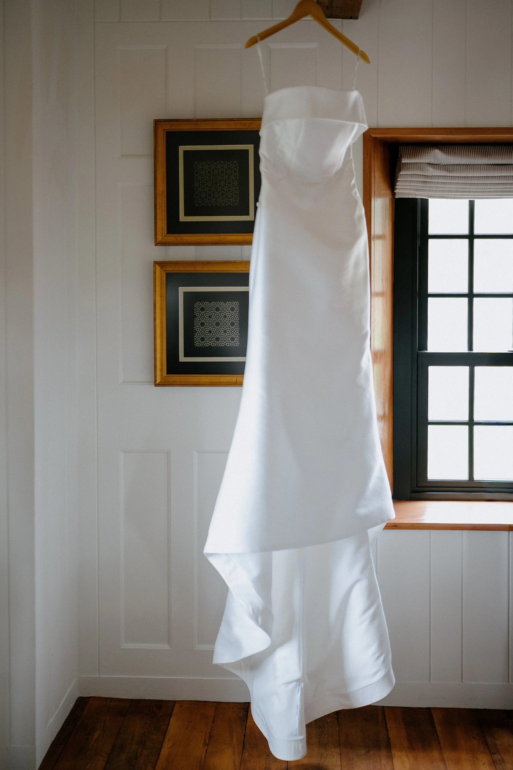 White wedding dress hanging on a wooden hanger in front of a window with wooden trim.