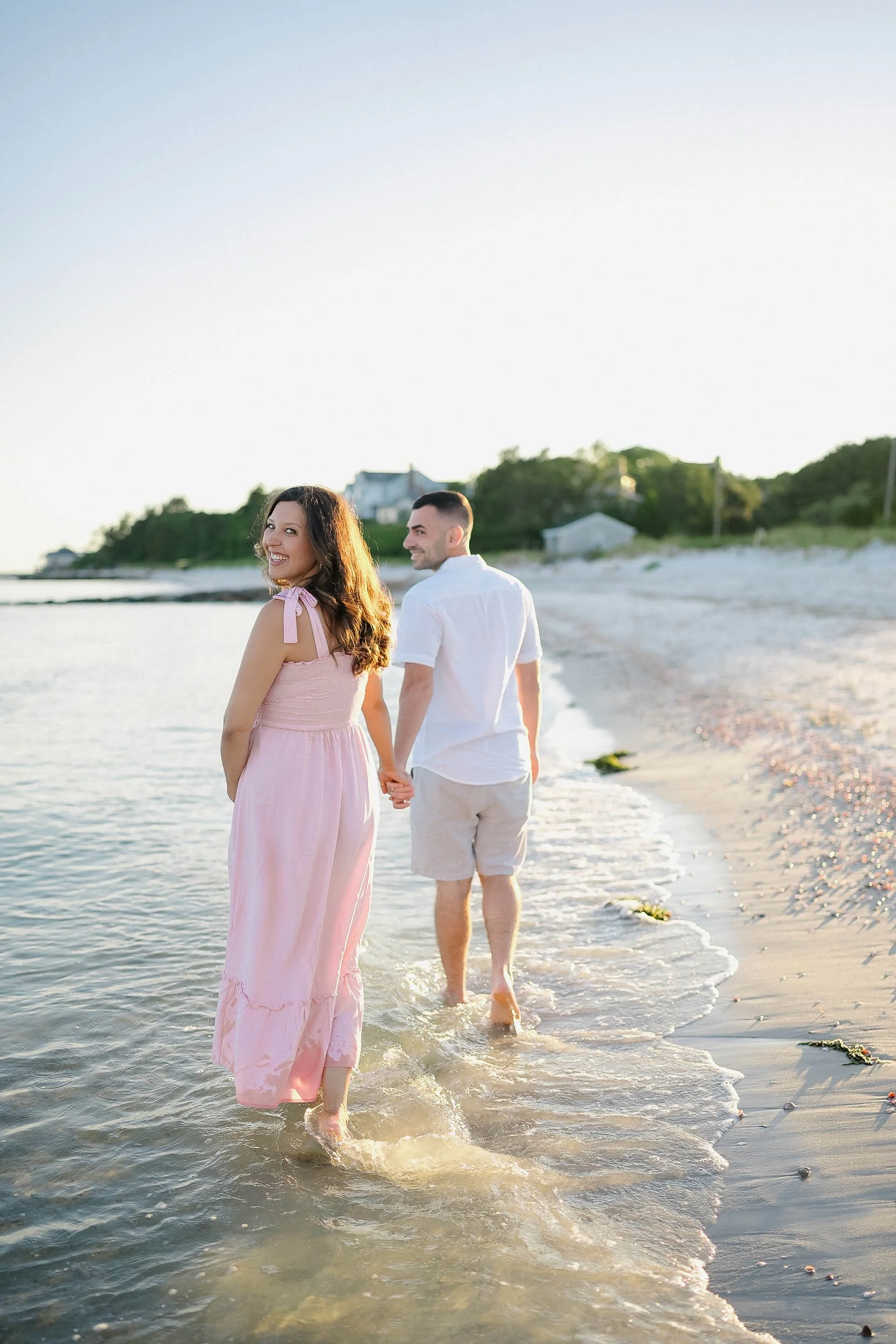 A couple walking hand in hand on the beach at sunset, with the woman wearing a pink dress and the man wearing a white shirt and beige shorts.