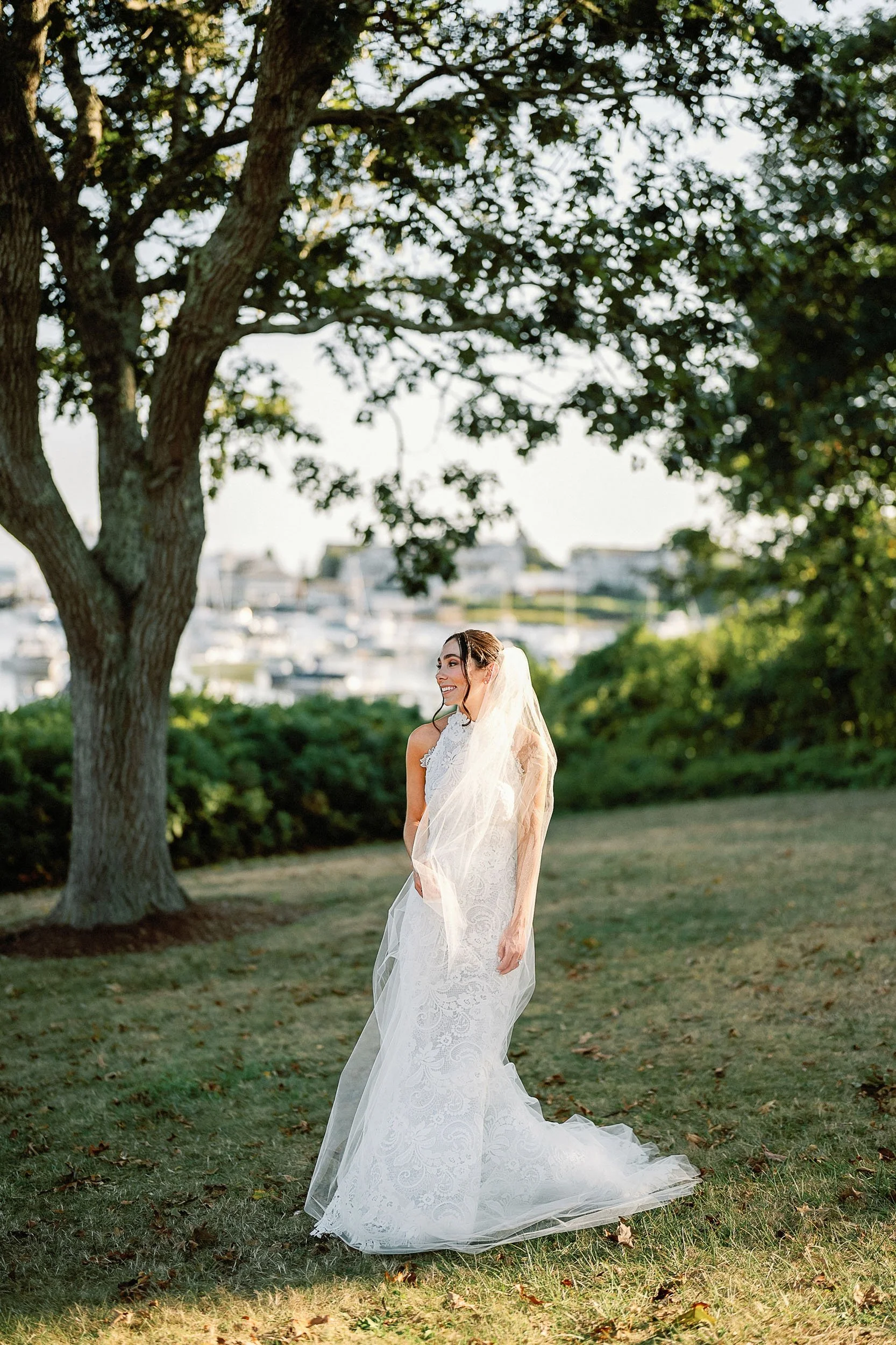 A bride standing on grass near a tree, wearing a white lace wedding dress with a veil, smiling, with boats docked at a marina visible in the background.