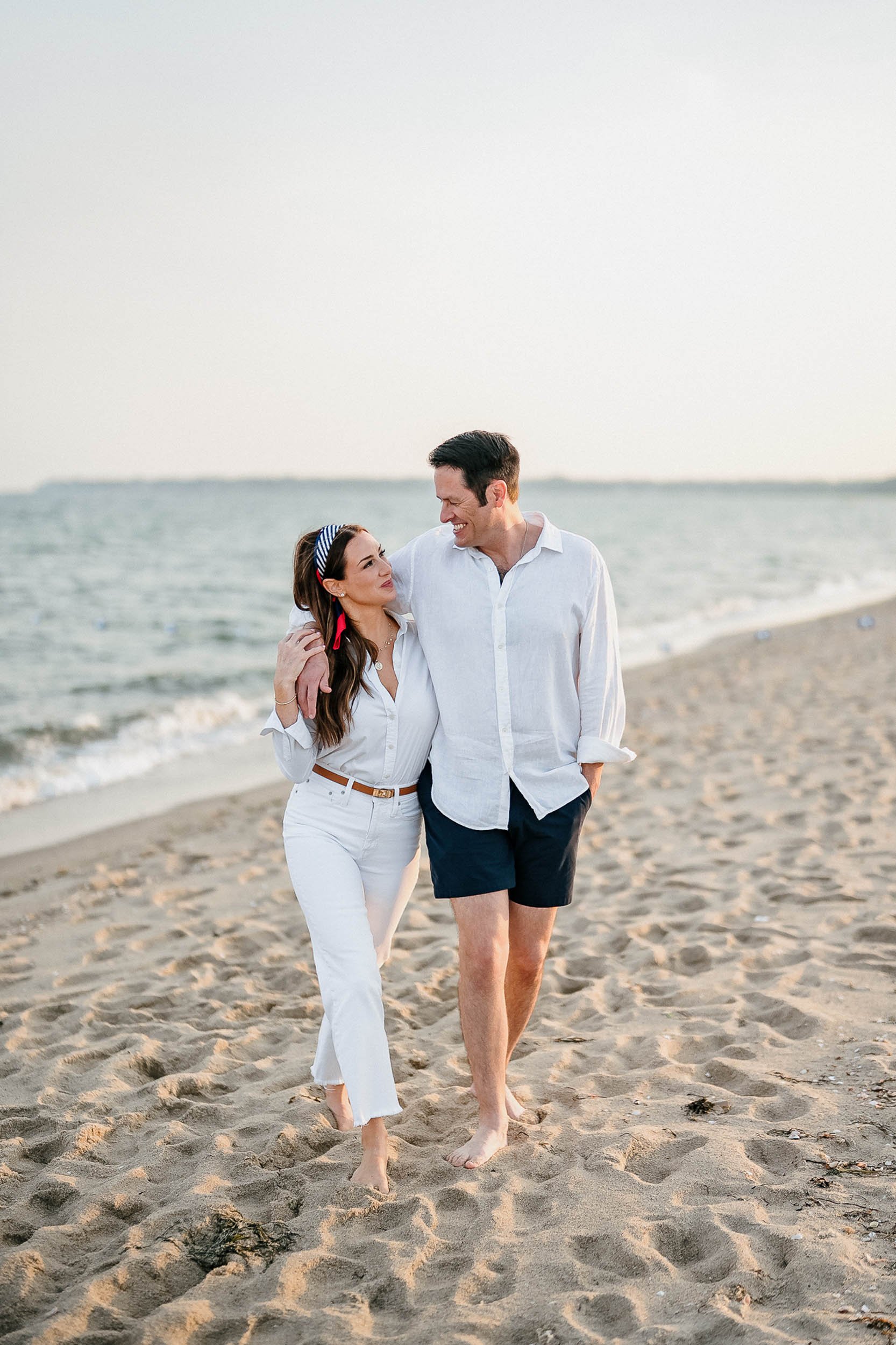 A couple walking on the beach, smiling and looking at each other, with the ocean and a cloudy sky in the background.