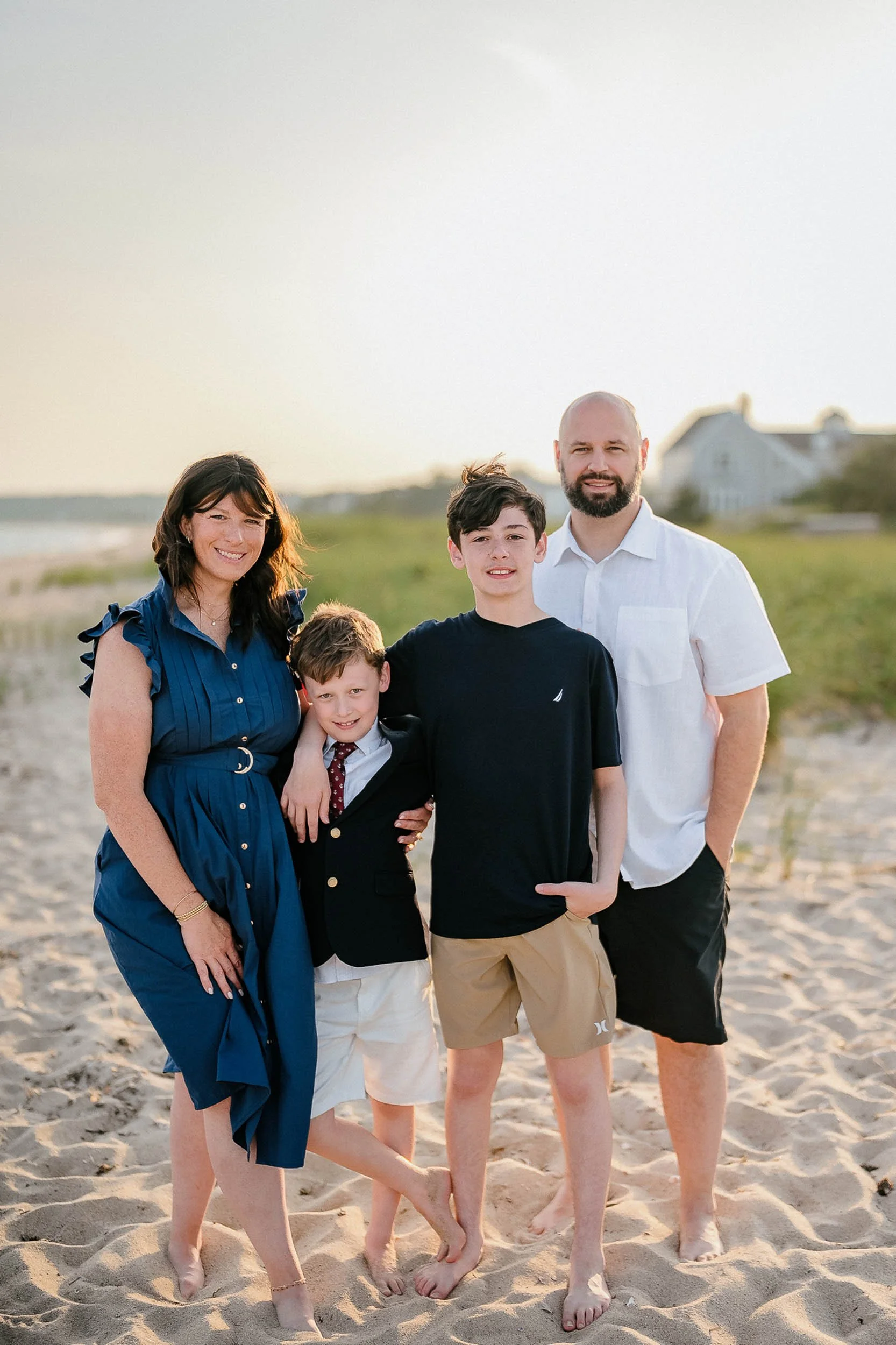 A family of five standing on a sandy beach during sunset, smiling at the camera. The mother is wearing a blue dress, the father is in a white shirt and shorts, and three boys are dressed in casual and semi-formal outfits.