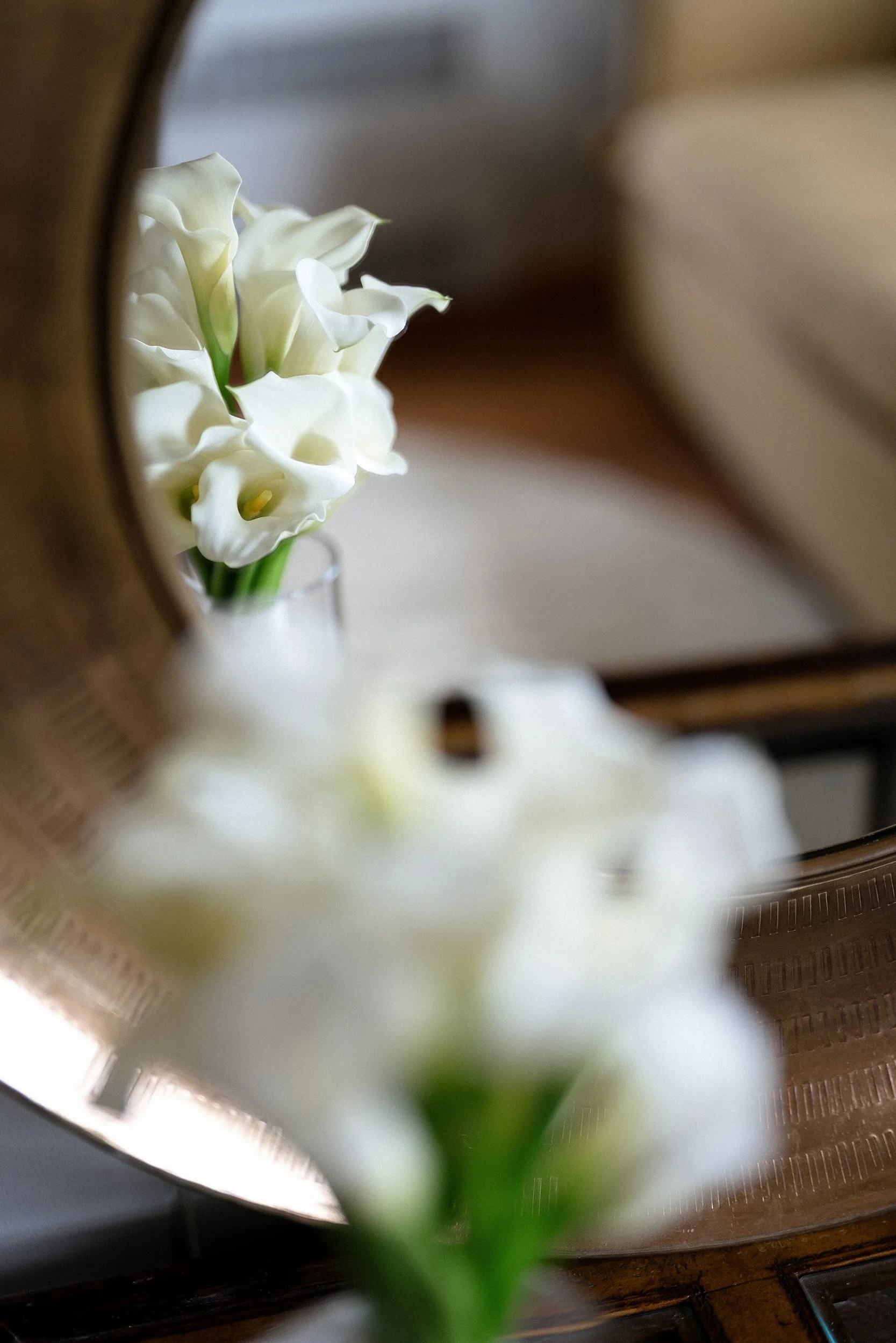 White flowers in a clear vase, reflected in a mirror with a brass frame, with blurred background.