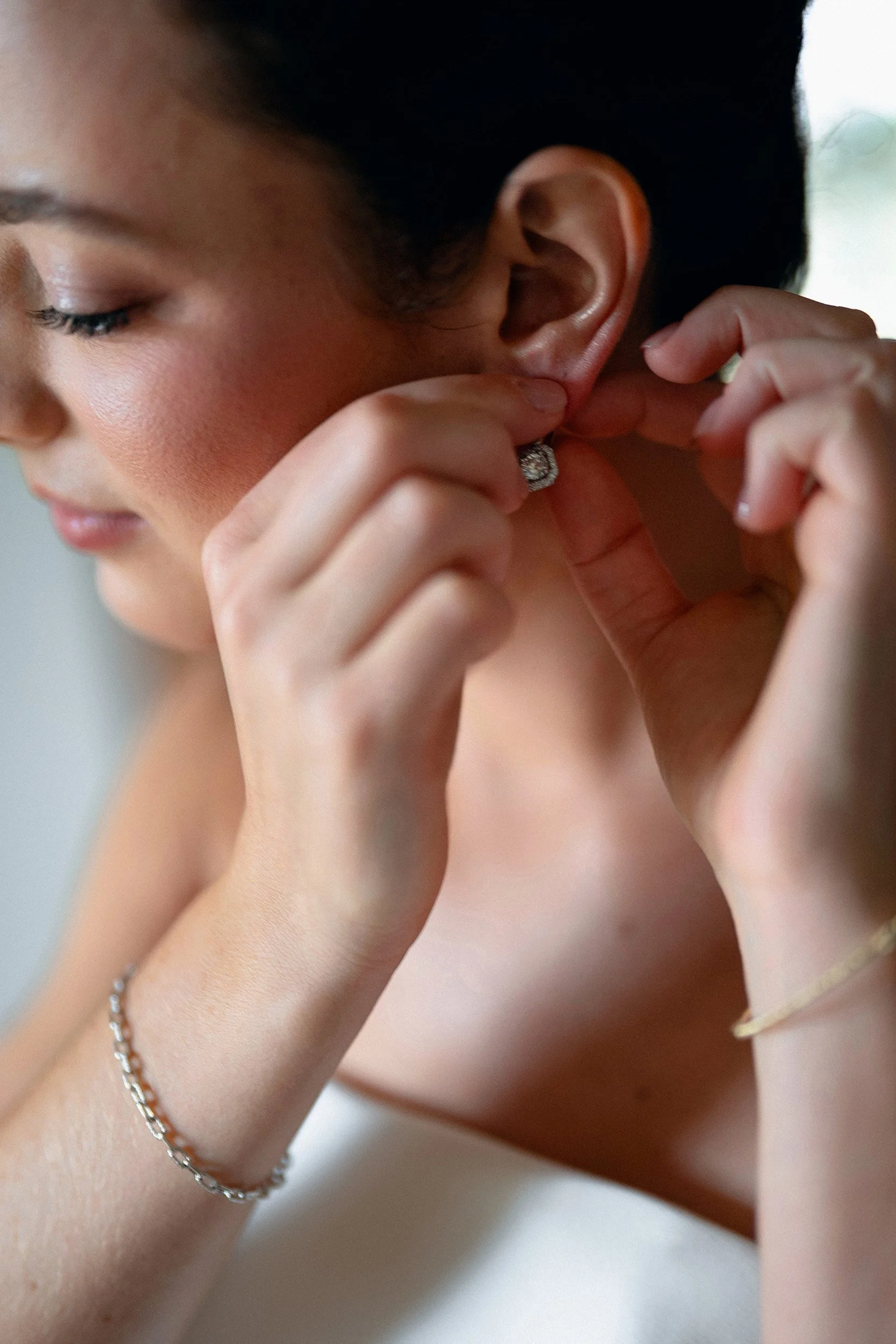 A woman with fair skin and dark hair is putting on a diamond earring. She is wearing a bracelet and a ring on her left hand.