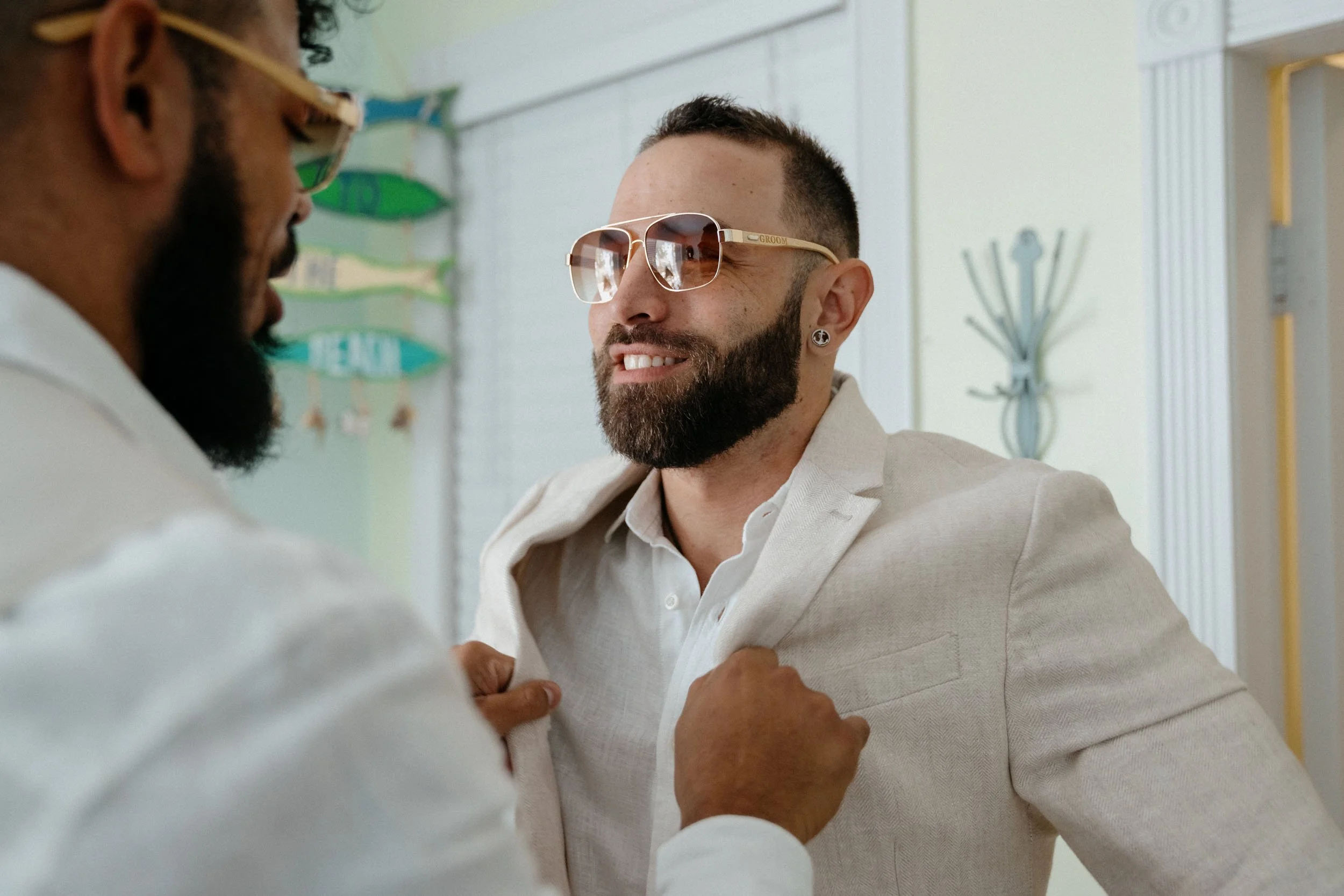 A groom in sunglasses smiling as a man pins a boutonniere onto his white suit jacket. The scene takes place in a brightly decorated room with hanging signs and a coat rack in the background.