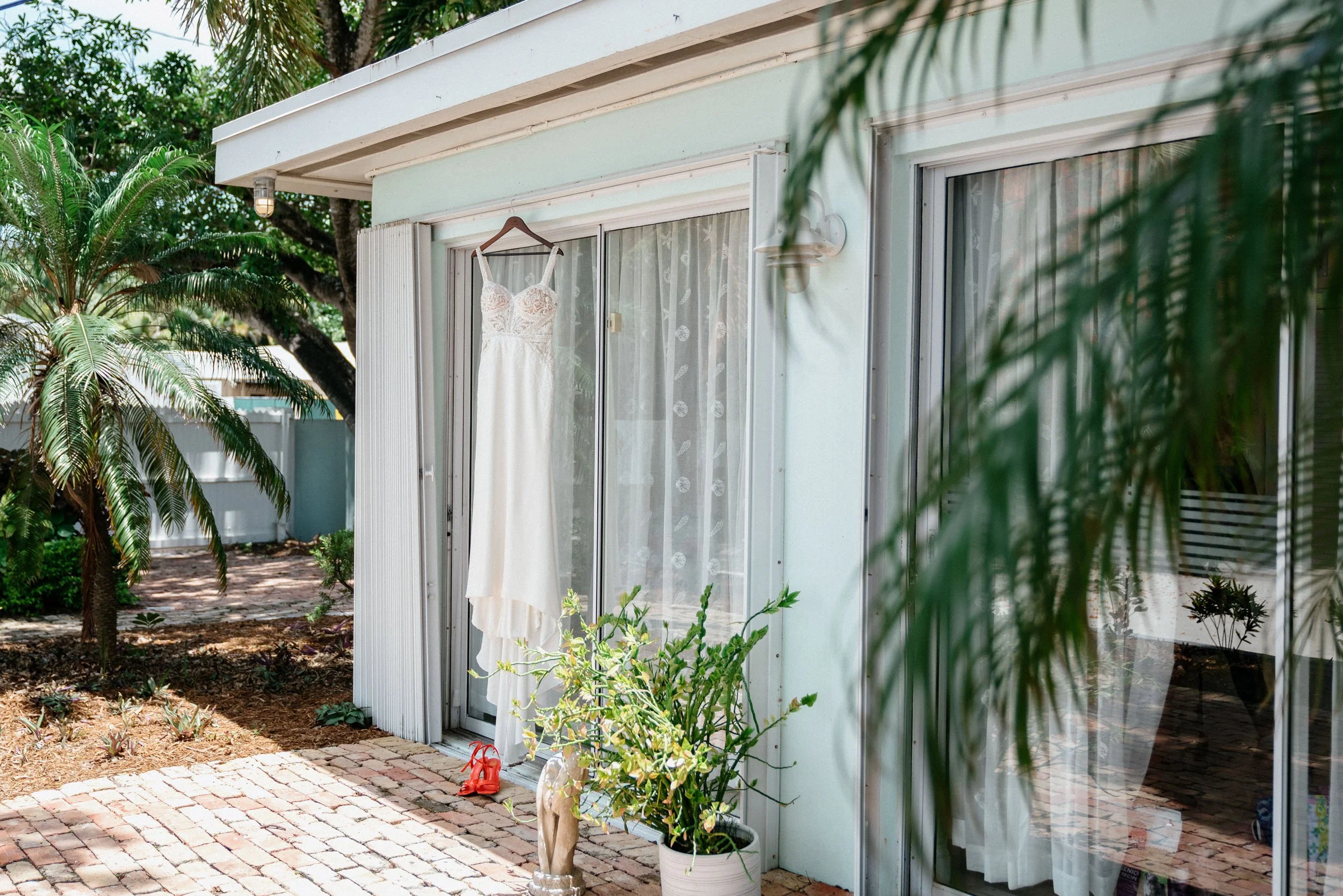 Wedding dress hanging on a hanger outside a house, with potted plants, brick patio, and sliding glass doors.