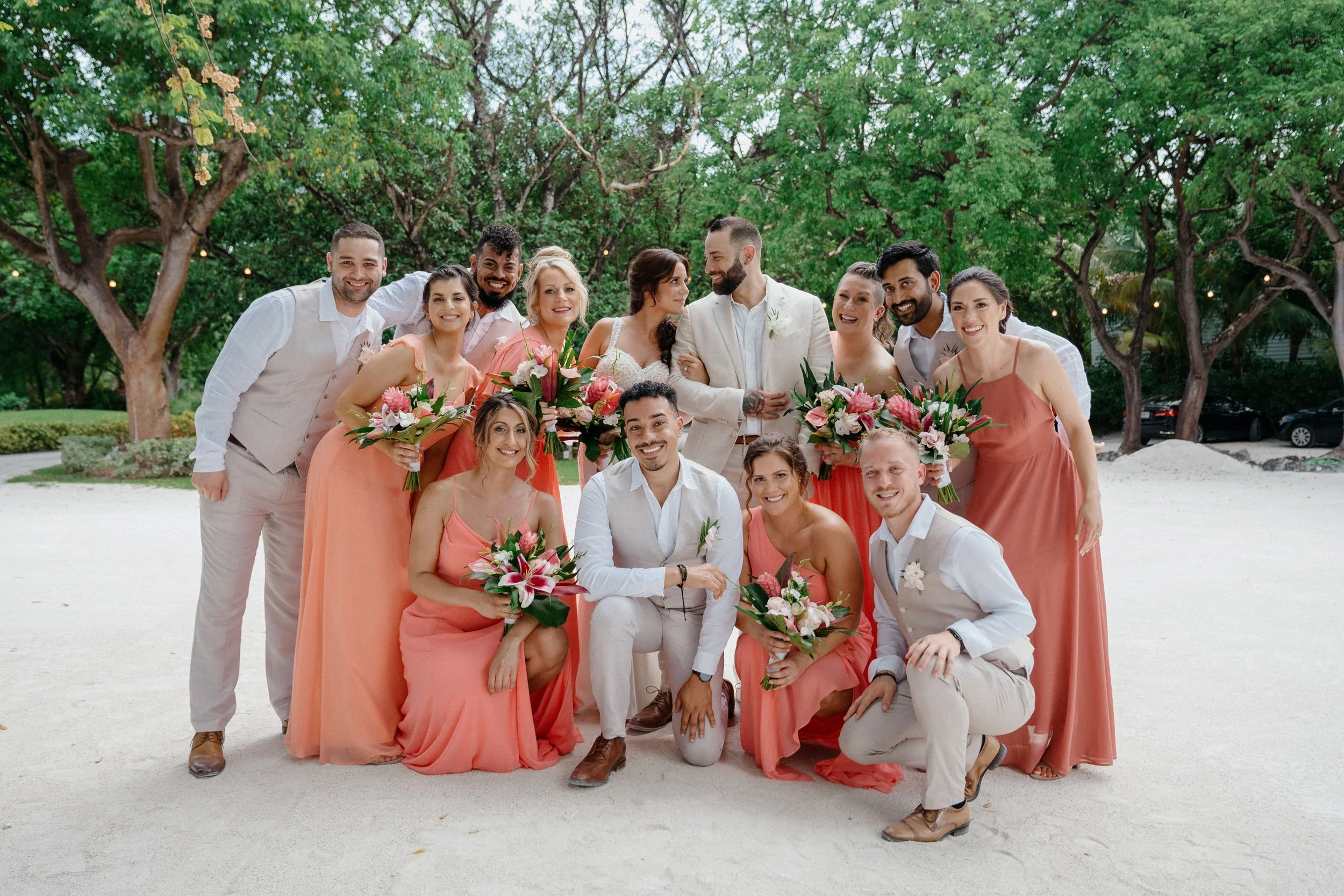 A wedding party outdoors on a sandy area with trees in the background, including the bride and groom, bridesmaids, and groomsmen. The women are wearing peach-colored dresses and holding bouquets, while the men are in light-colored suits or shirts. Ev