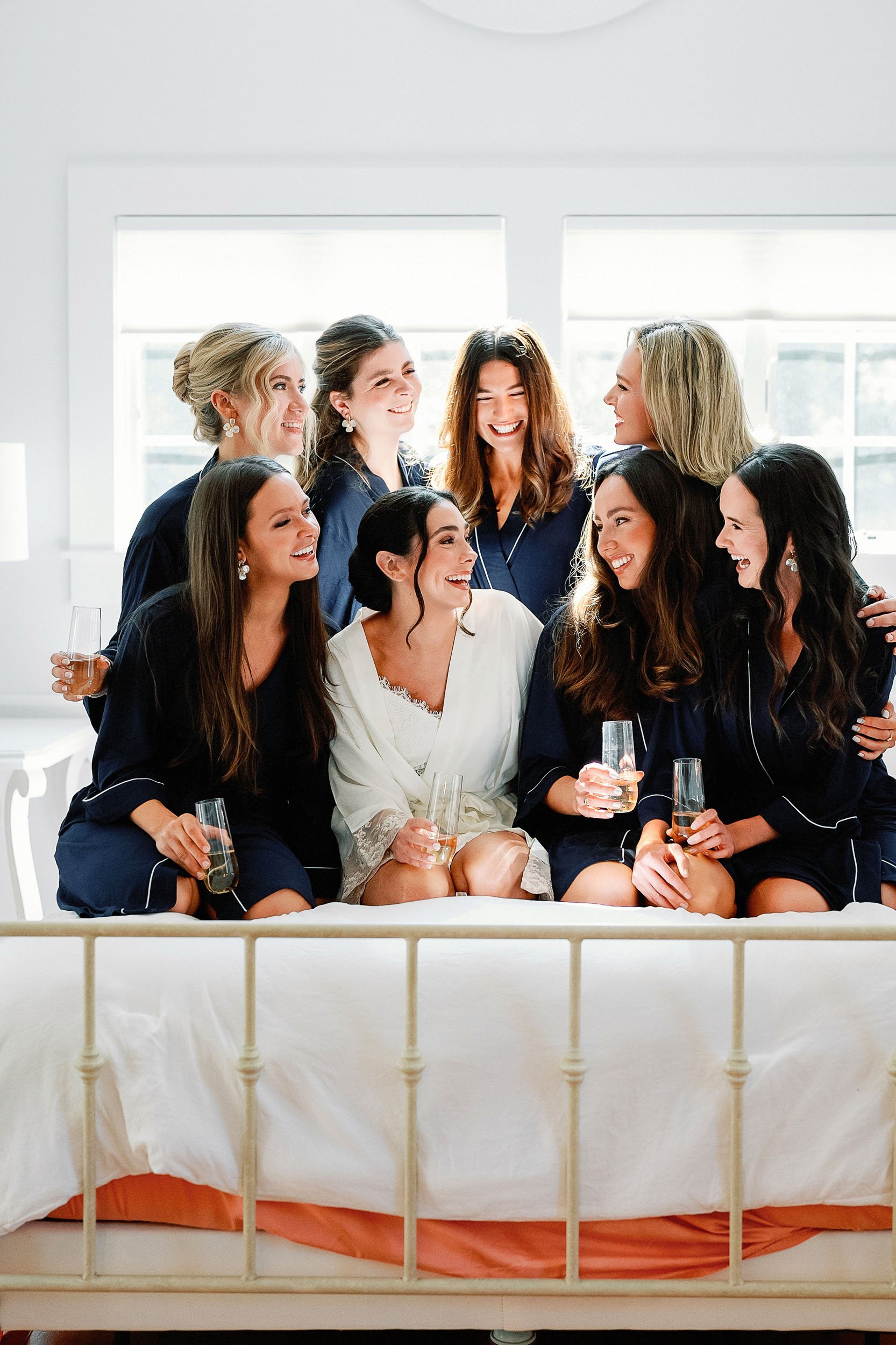 A group of women, dressed in matching navy blue pajamas, sitting on a bed, celebrating with champagne. They are smiling and laughing, with sunlight streaming through the windows behind them.