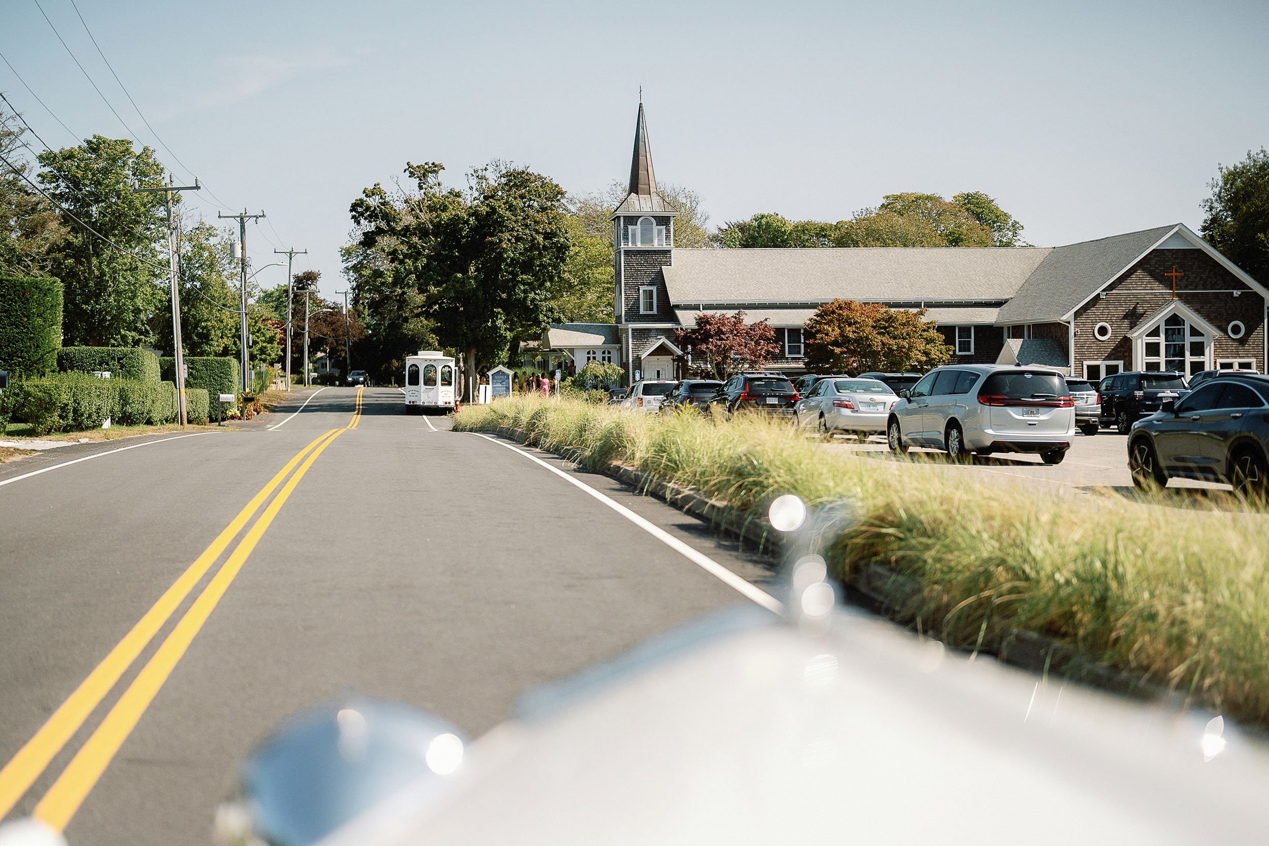 A street scene with a church, parked cars, a small white bus, and trees, viewed from inside a car on a sunny day.