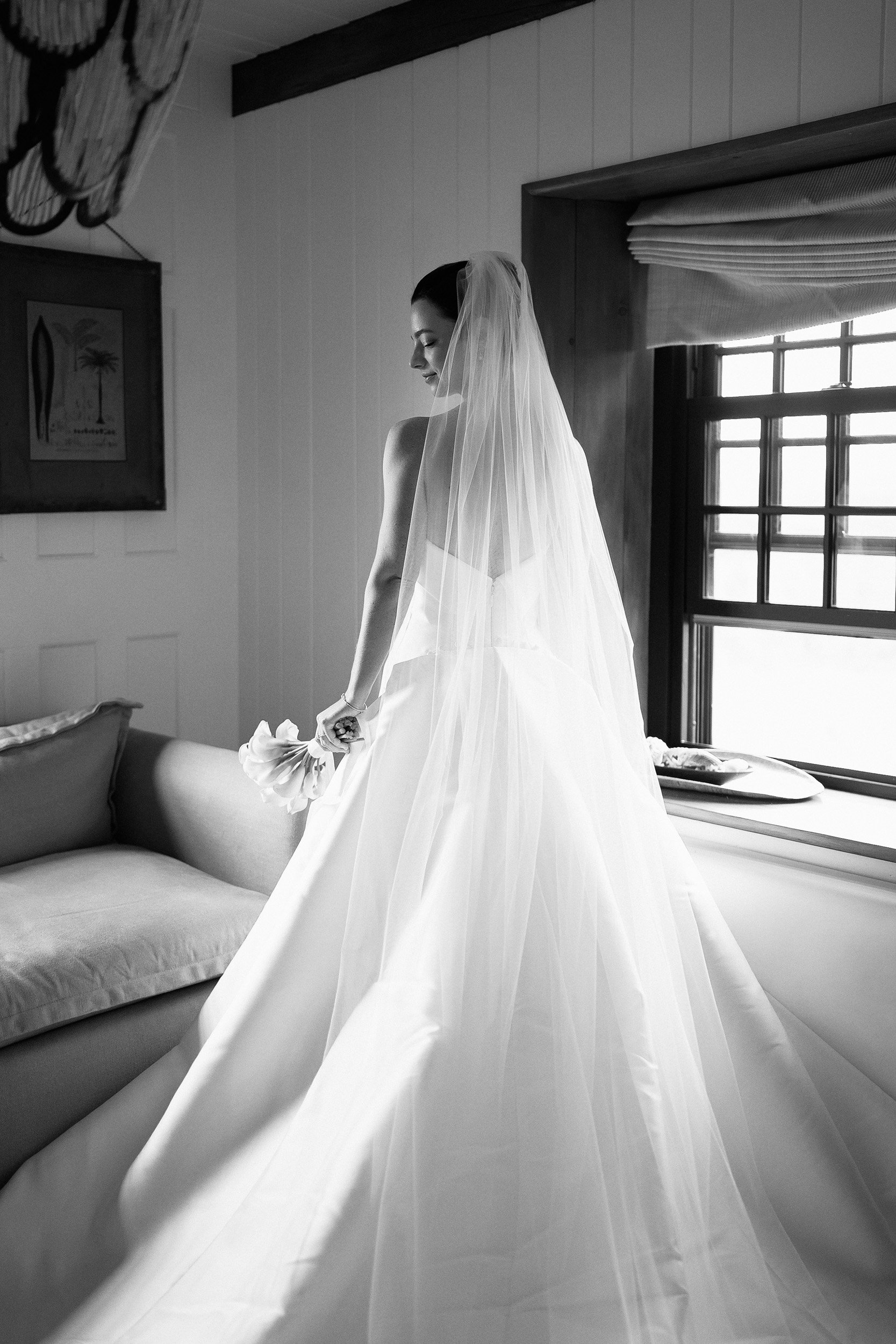 Black and white photo of a bride in a wedding dress with a veil holding her bouquet, standing by a window