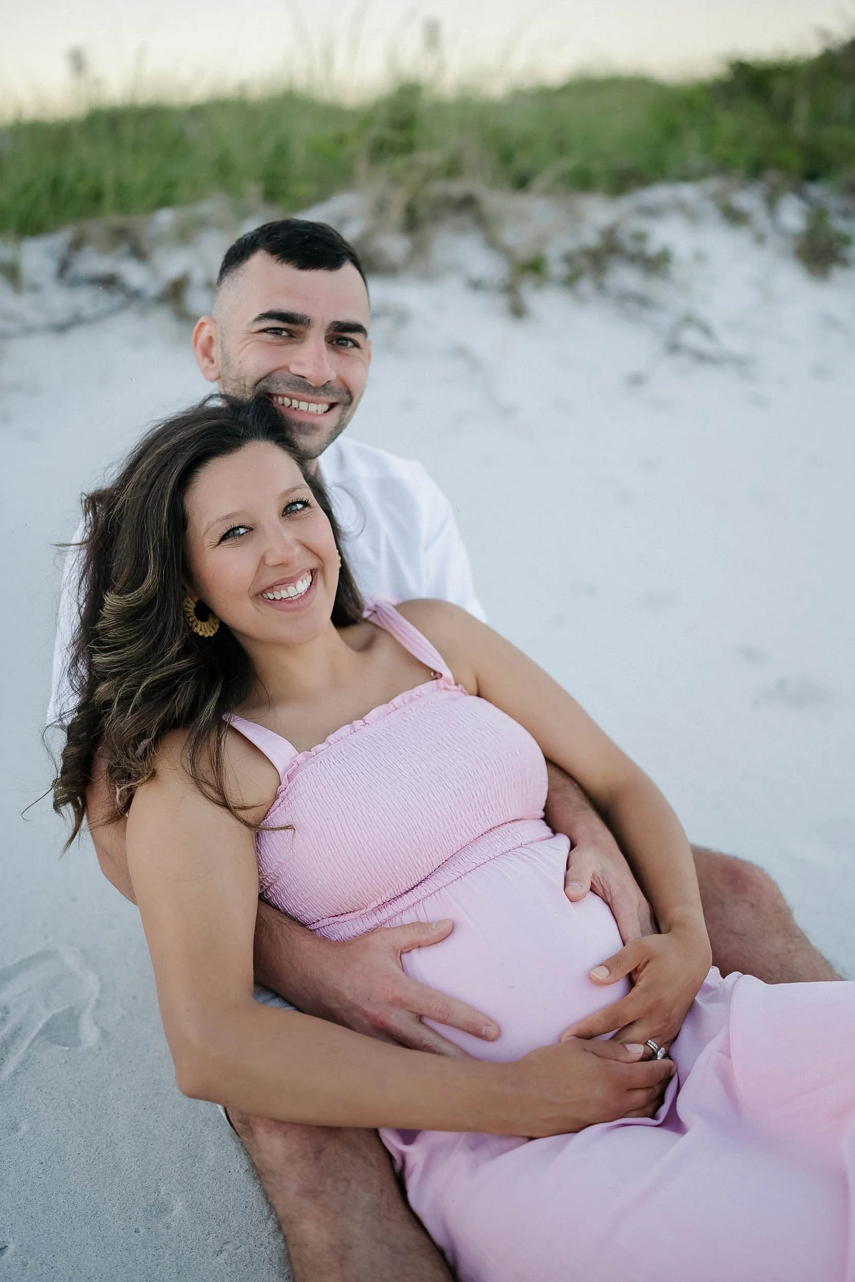 Happy couple sitting on the sand at the beach, with the man holding the pregnant woman.