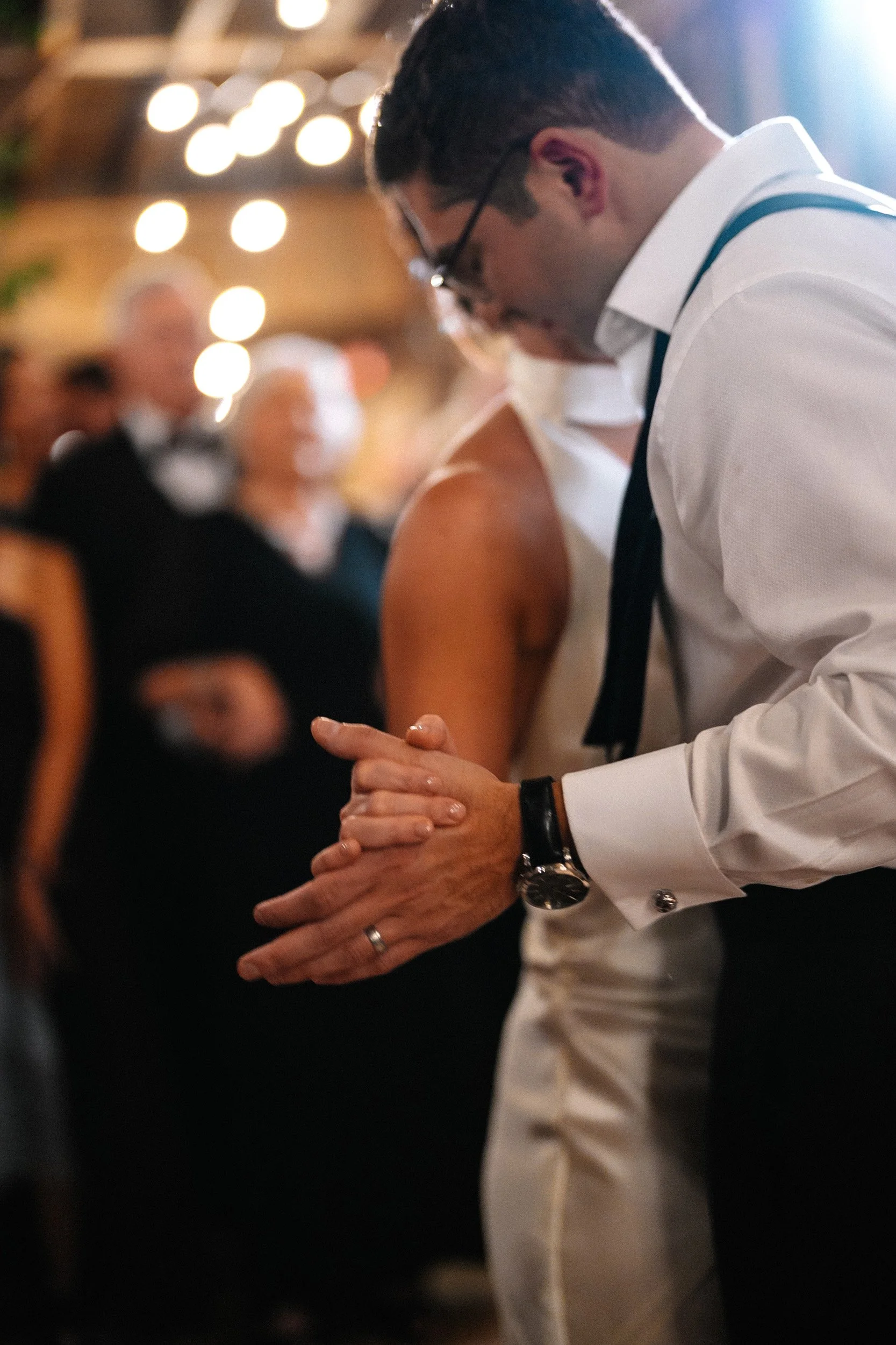 A man and woman dancing closely at a formal event, with the man dressed in a white shirt and black tie, and the woman in a sleeveless dress. The background shows an audience of formally dressed people and blurry lights.