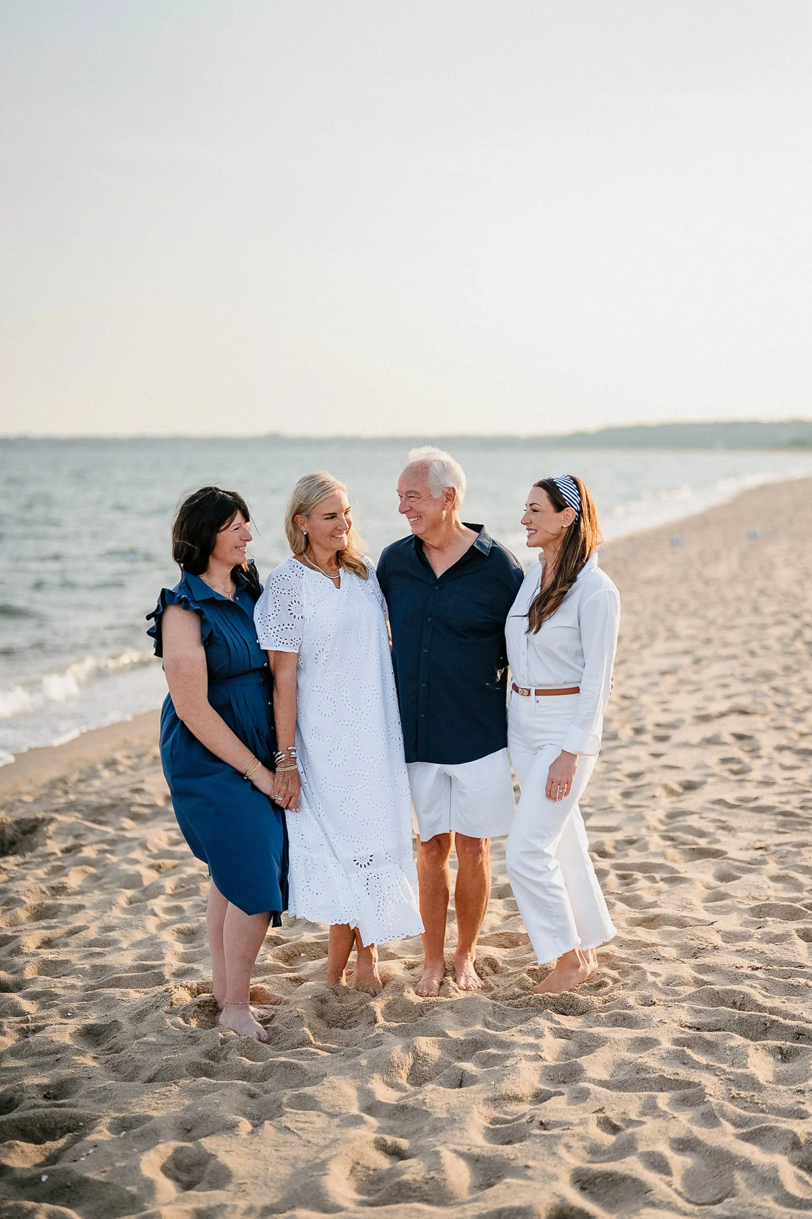 Four people standing on a sandy beach near the water, smiling and enjoying each other's company during sunset or late afternoon.