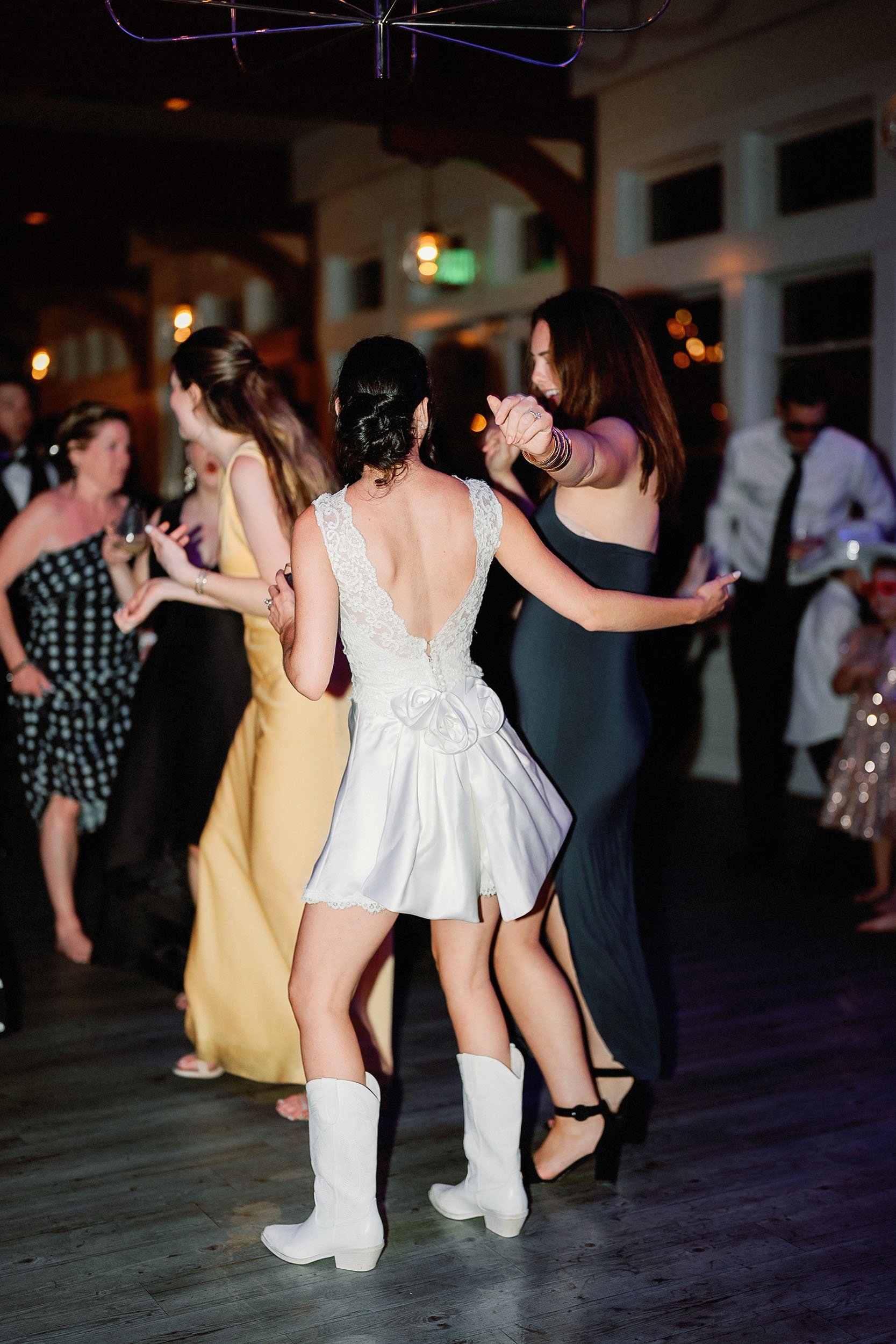 People dancing at a party, with one woman in a white dress and cowboy boots, and another woman in a black dress, in a warmly lit indoor setting.