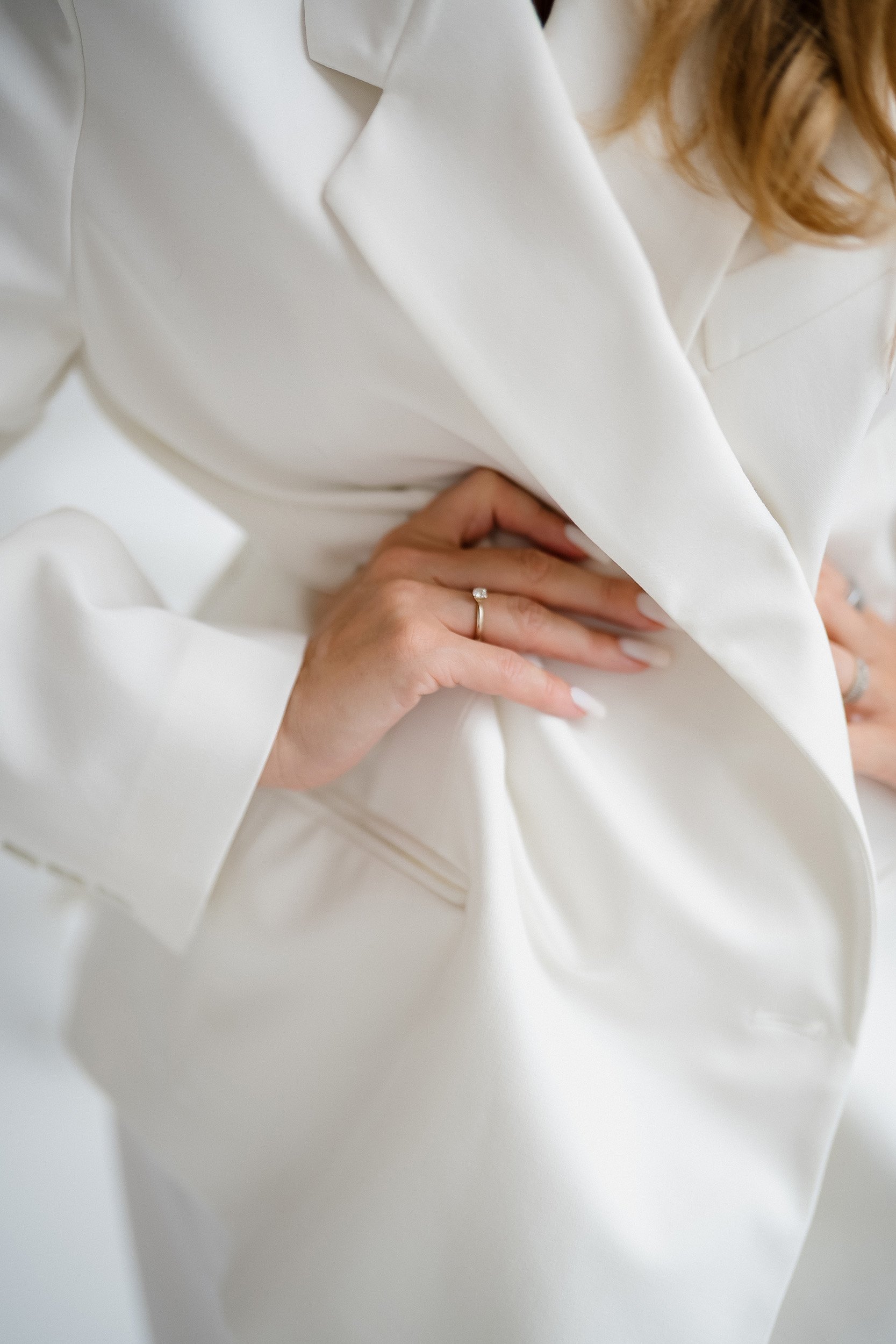 Close-up of a person wearing a white jacket, with their hand resting on their waist, showing a ring with a small gemstone.