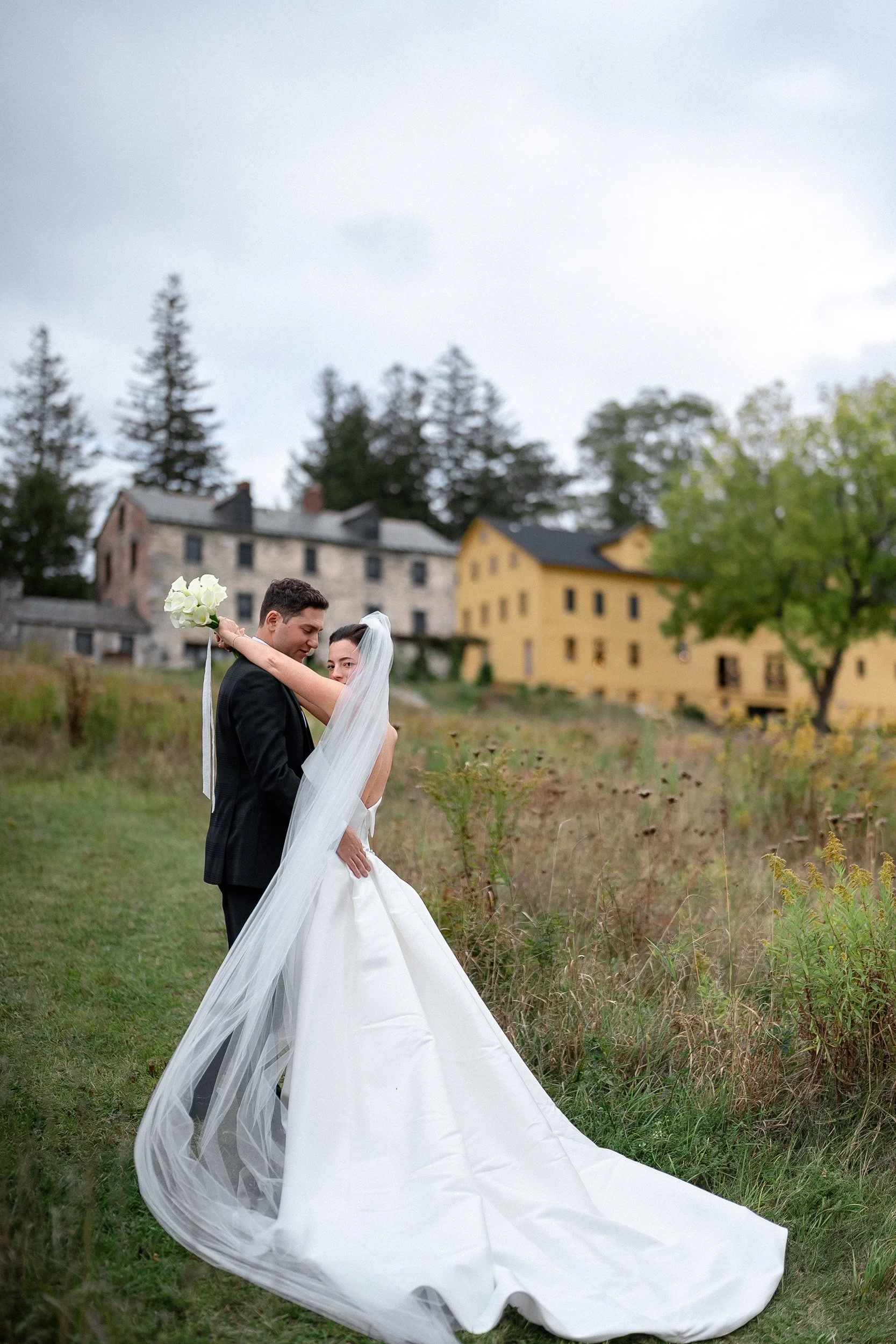 A bride and groom embrace outdoors on a cloudy day, with the groom holding a bouquet of white flowers. The bride wears a long white wedding gown and veil, and the groom is in a black suit. There are trees and buildings in the background.