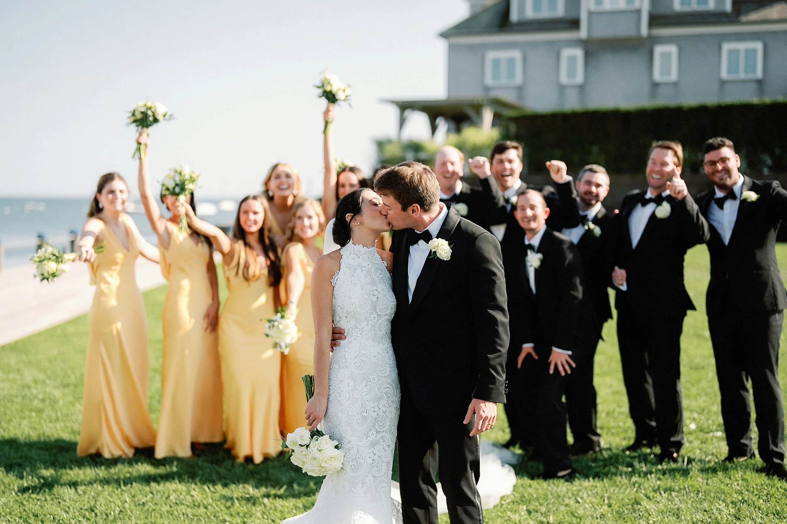 Bride and groom kissing in front of wedding party on a grassy outdoor area near the water, with bridesmaids in yellow dresses and groomsmen in black tuxedos, some holding bouquets, celebrating.