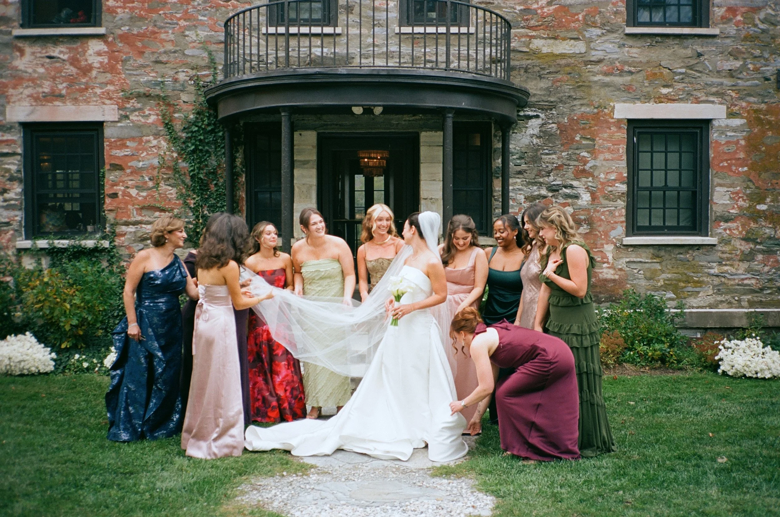 A bride with a white dress and veil surrounded by a group of women in fancy dresses, some of whom are assisting with her wedding gown outside a stone building with black framed windows and a black door.
