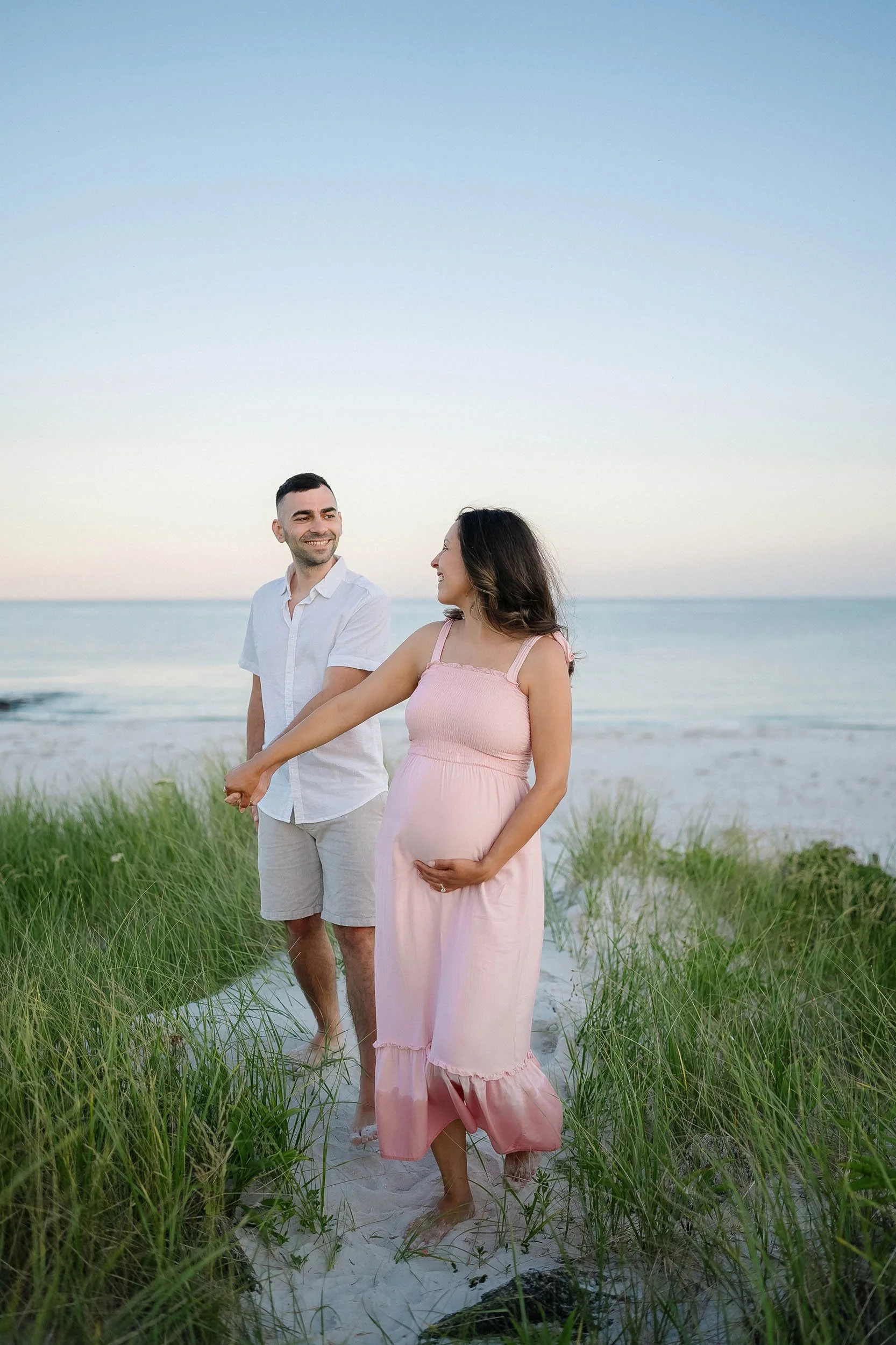 A pregnant woman in a pink dress holding her belly walks hand in hand with a man on a beach with green grass, sand, and ocean in the background during sunset or sunrise.