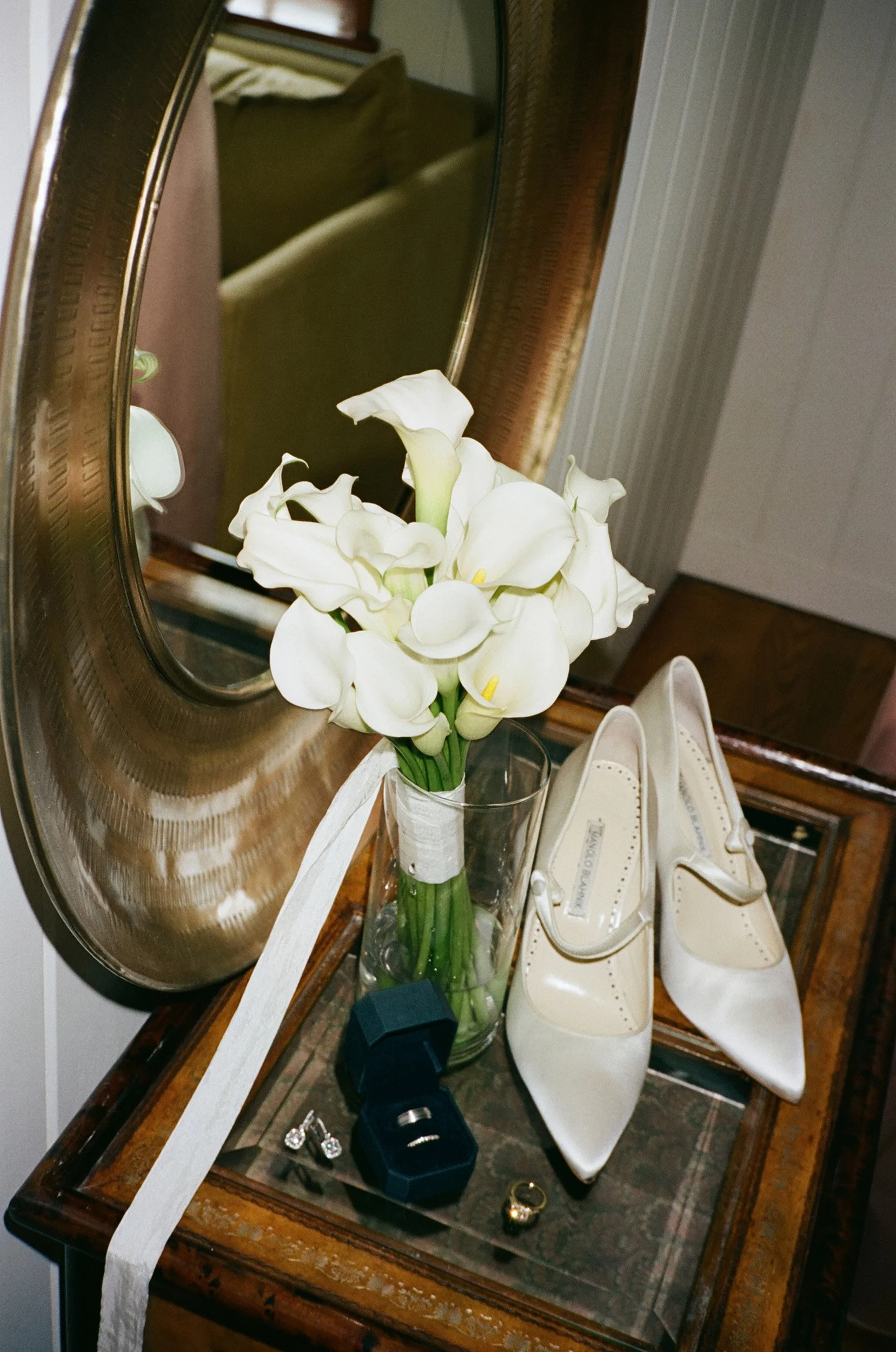 A vintage wooden dressing table with a large mirror, holding white calla lilies in a glass vase, a pair of white high-heeled shoes, an open jewelry box with rings, and a pair of earrings.