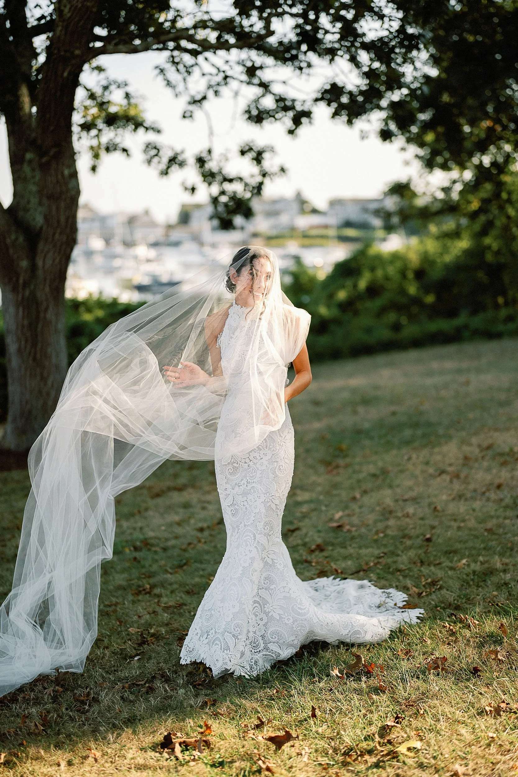 Bride in a white lace wedding gown with a long train standing outdoors near a tree, wearing a veil and looking off to the side with a marina and boats in the blurred background.
