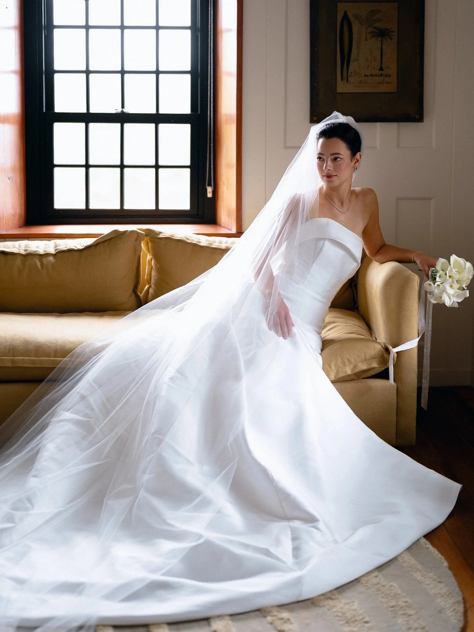 A bride in a white wedding dress and veil sitting on a beige sofa, holding a bouquet of white calla lilies, with a large window behind her and framed artwork on the wall.