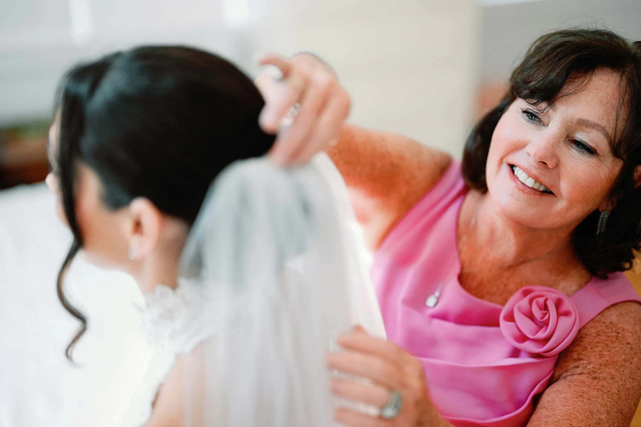 A woman in a pink dress smiling as she adjusts a bride's veil.