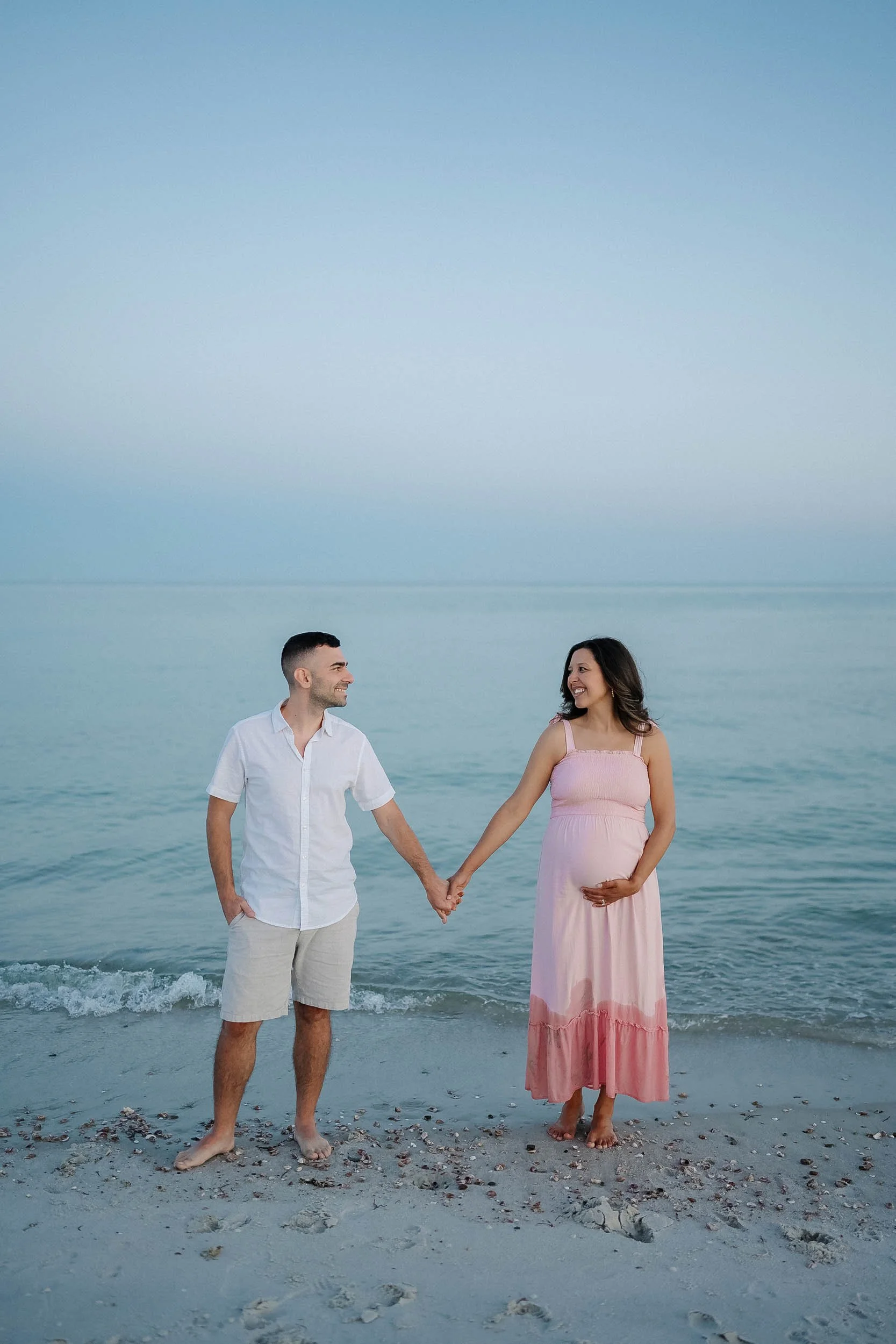 A pregnant woman in a pink dress holding hands with a man in a white shirt and beige shorts on the beach during sunset.