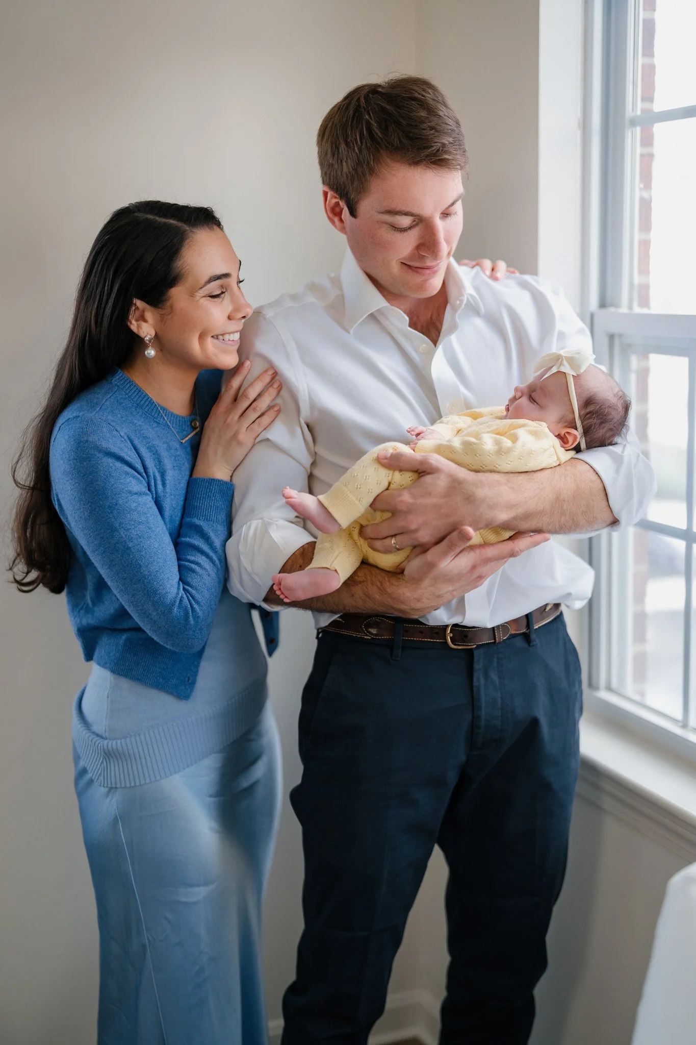 A young couple holding a sleeping baby girl near a window, with the mother smiling and touching the father's shoulder.