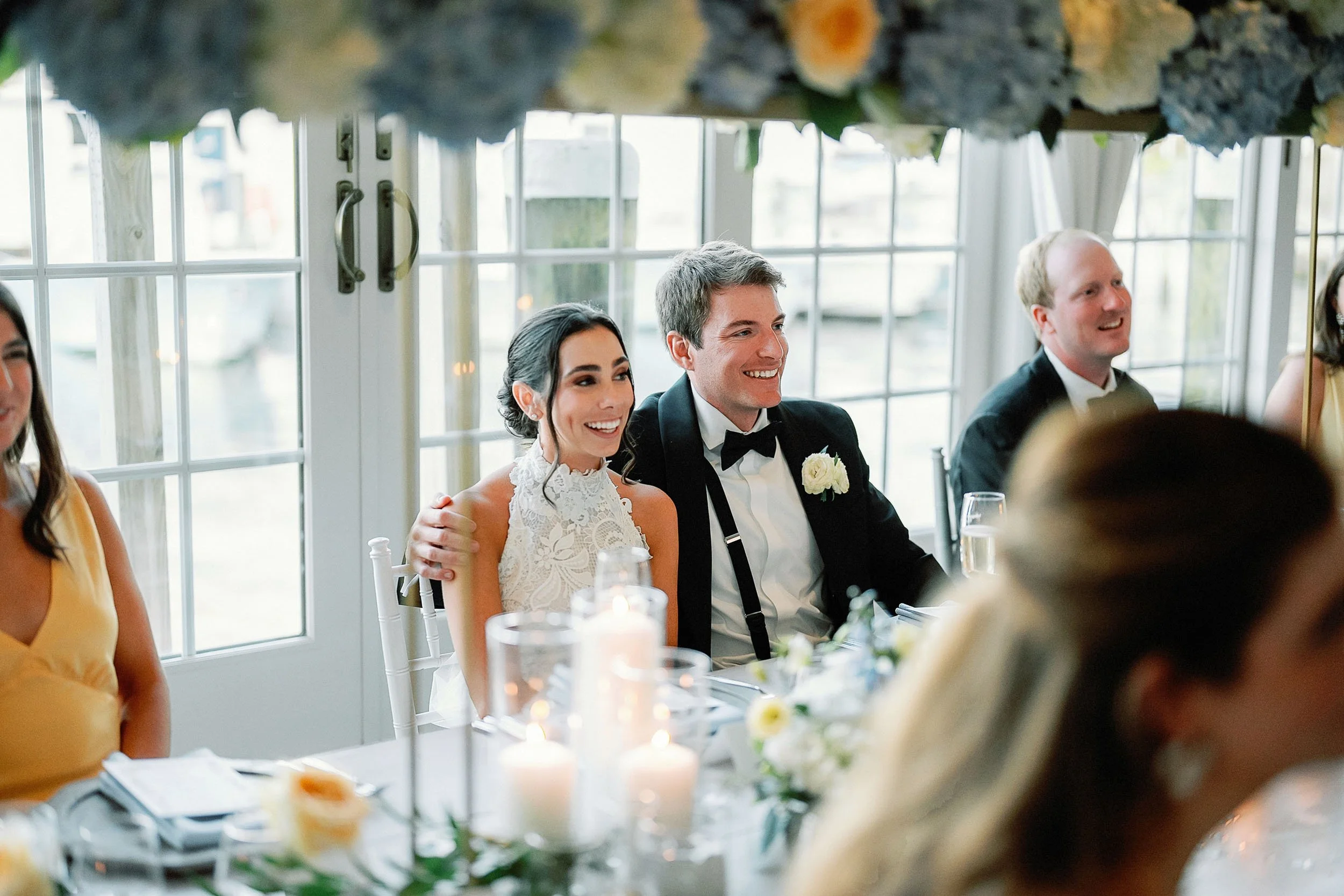 Bride and groom sitting at a wedding reception table smiling, with candles and flowers, near large windows with a view of boats on water.