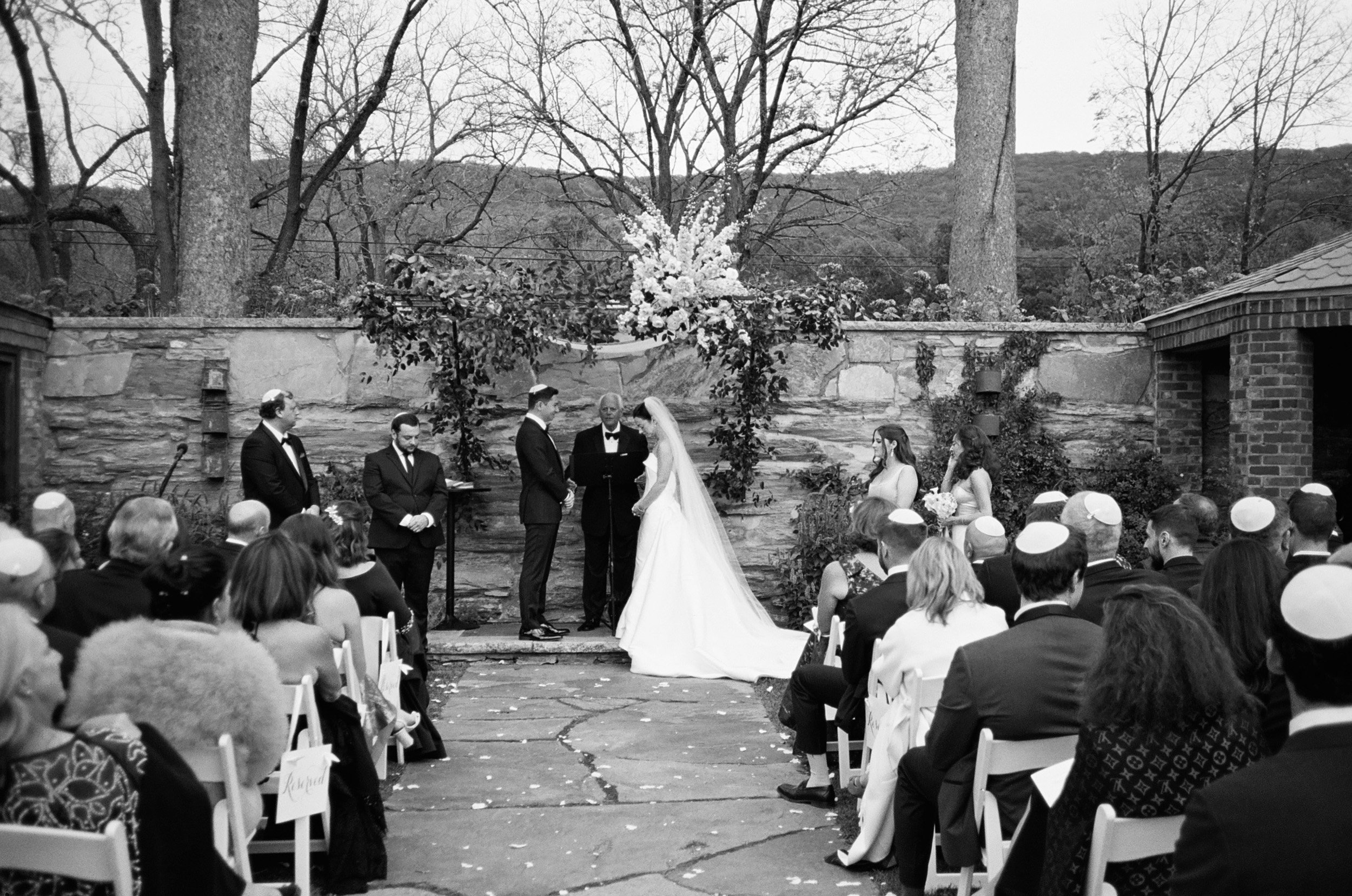 A black and white photograph of a wedding ceremony outdoors with guests seated, the bride and groom holding hands at the altar, surrounded by bridesmaids and groomsmen, under an arch decorated with flowers and greenery.