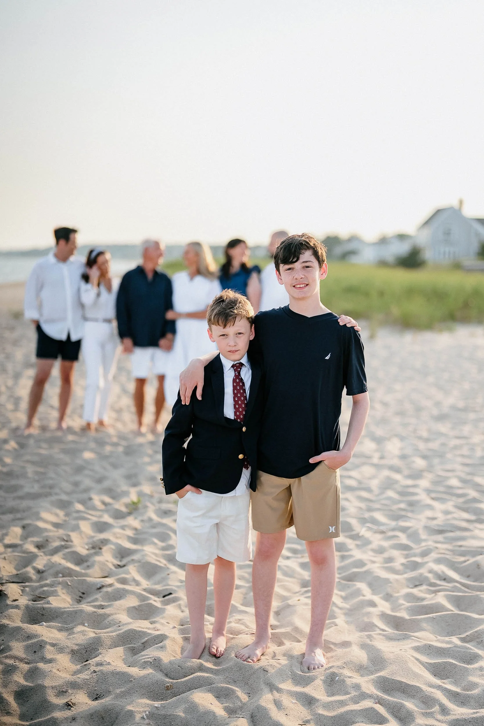 Two boys standing barefoot on the beach with an out-of-focus group of people in the background, possibly family, during sunset.