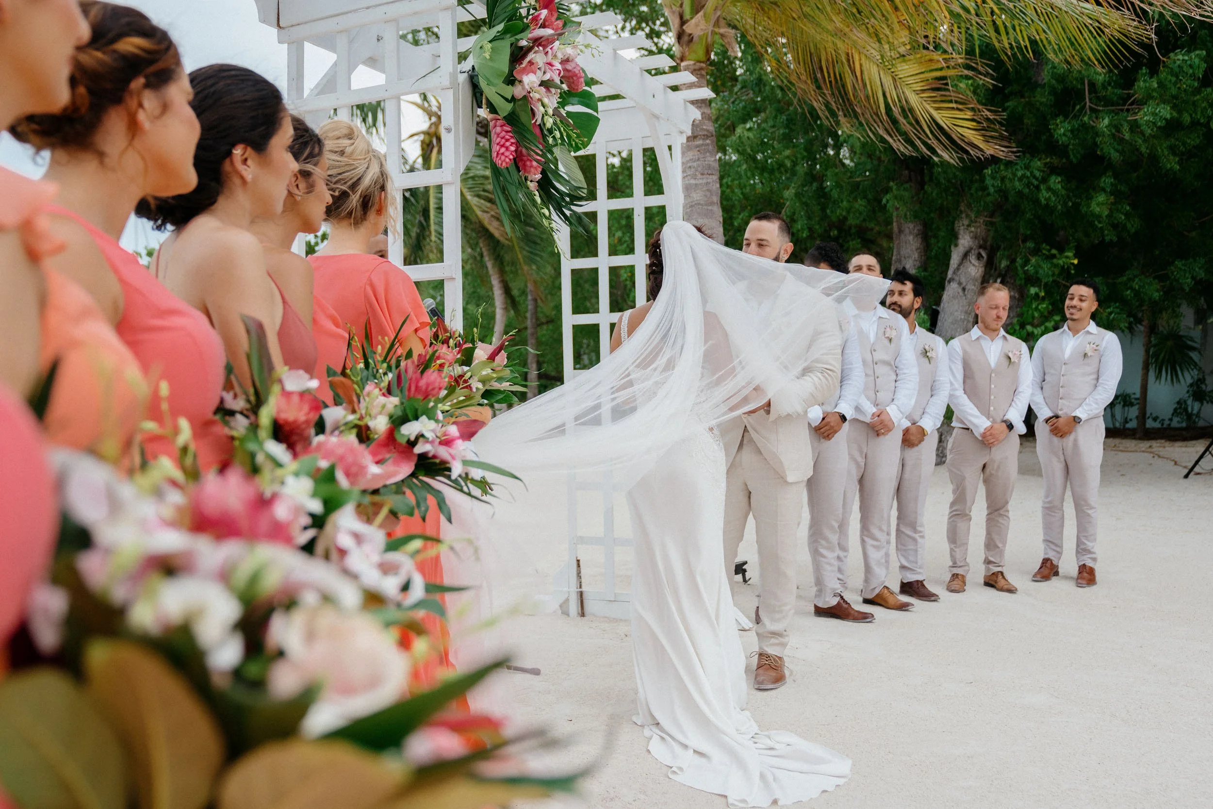 A wedding ceremony at the beach with the bride and groom standing under a white floral arch while the bridesmaids and groomsmen look on. The bride is wearing a wedding dress with a long veil, and the bridesmaids are in coral dresses holding bouquets.