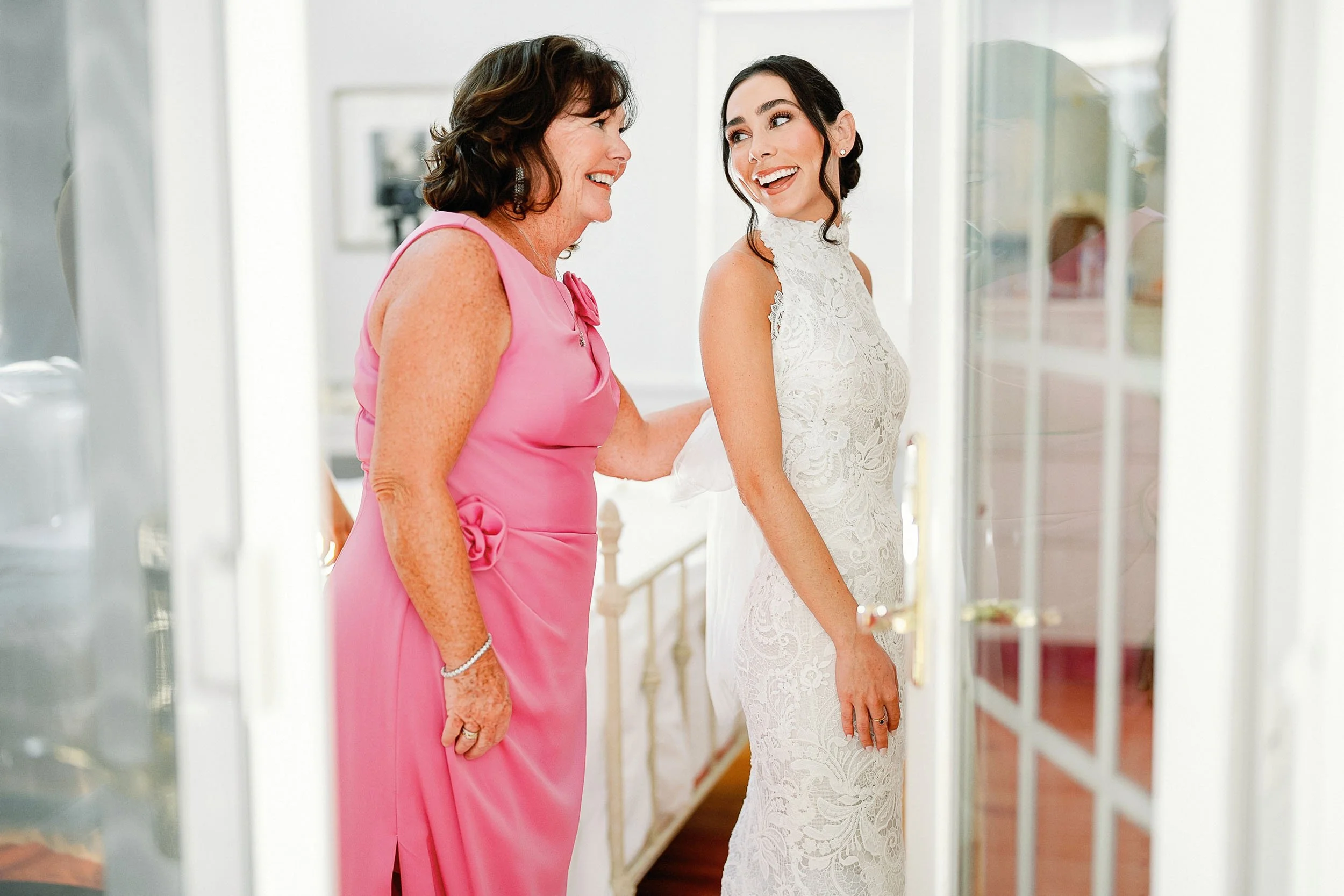 A woman in a white lace wedding dress smiling and looking at another woman in a pink dress, both standing inside a house, with the woman in the pink dress reaching out to the bride.
