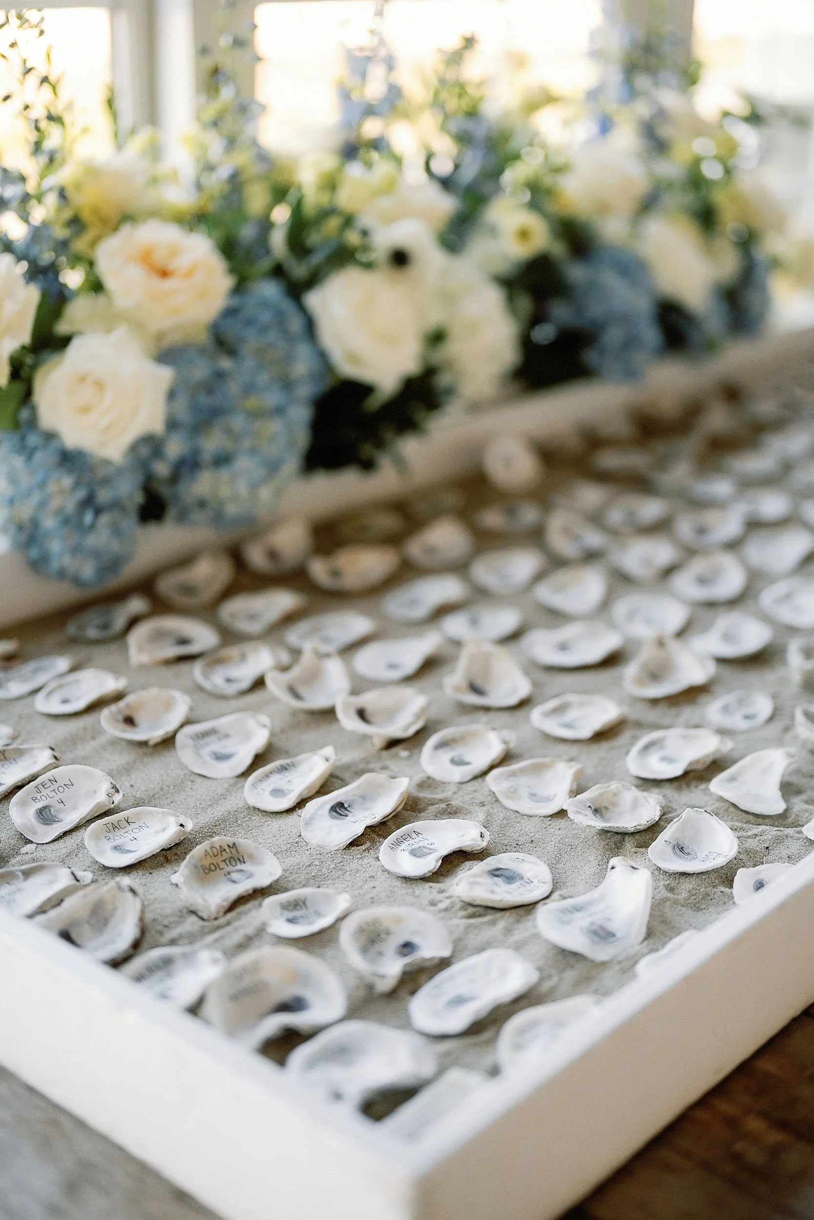 A tray filled with small paper shells, each with a handwritten name and eye drawn on them, likely for a wedding or event seating arrangement. In the background, there is a floral arrangement with white and blue flowers.