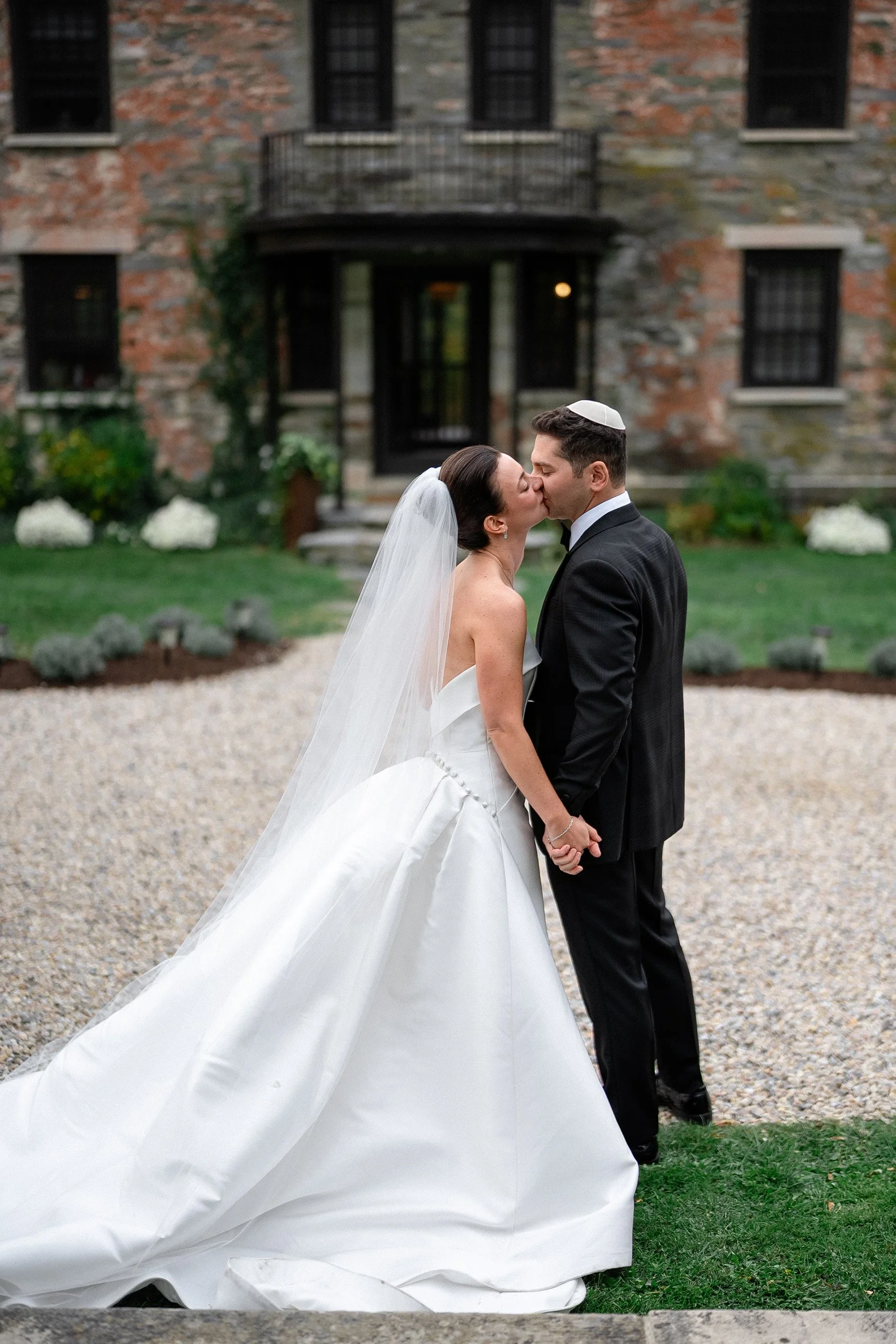 A bride and groom kissing outdoors in front of a brick and stone building, holding hands, with the bride in a white wedding gown and veil, and the groom in a black suit and yarmulke.