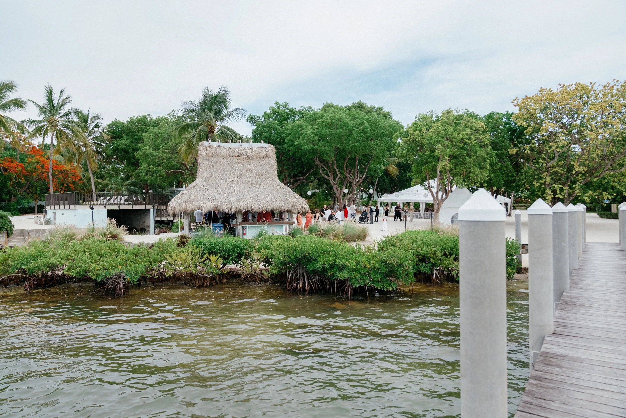A scene of a tropical outdoor area with palm trees, green bushes, and a thatched-roof hut by the water. There are white tents and people gathered in the background, and a wooden dock runs along the right edge of the image.
