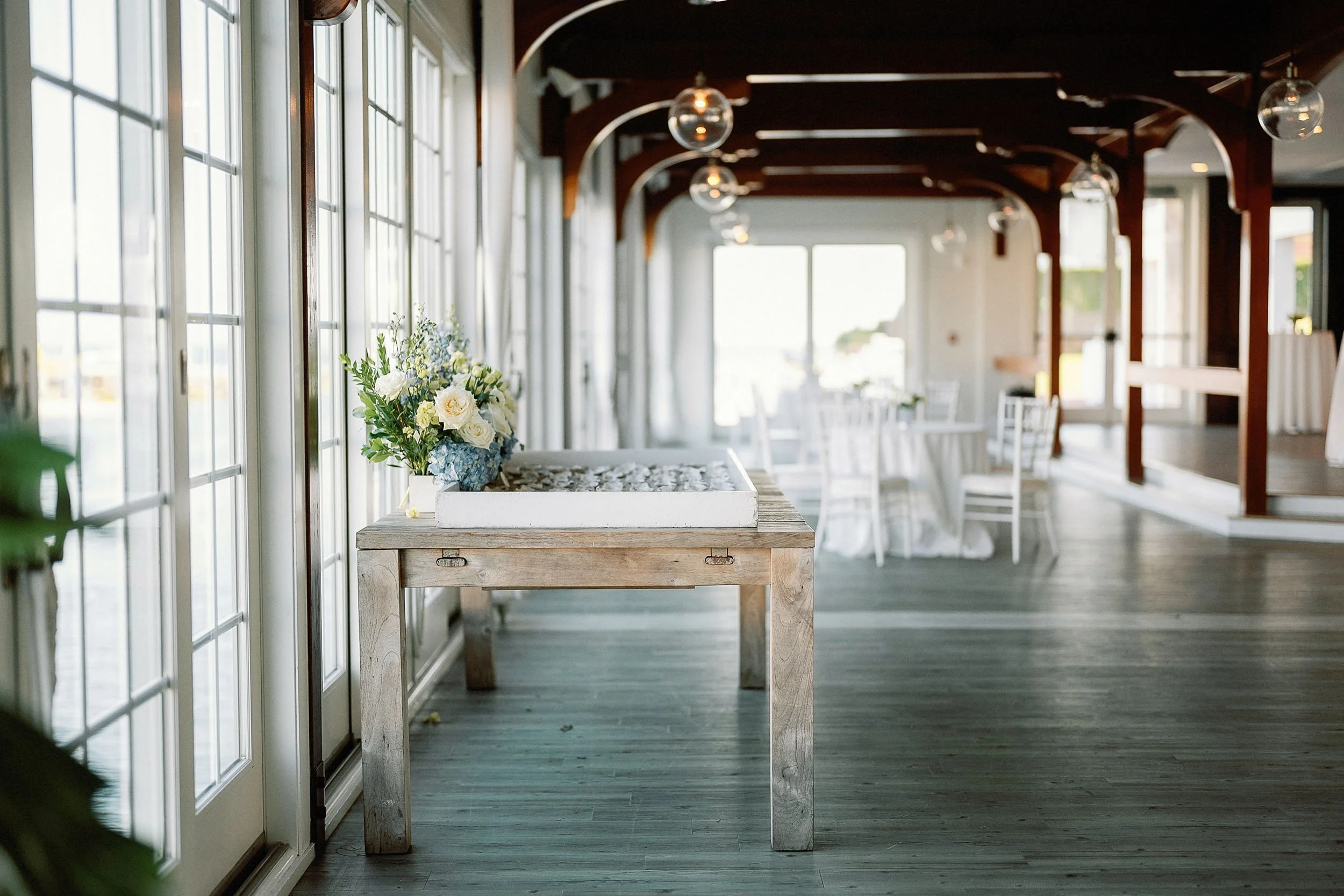 Empty event hall with large windows, wooden beams, and round tables with white tablecloths, decorated with floral centerpieces.