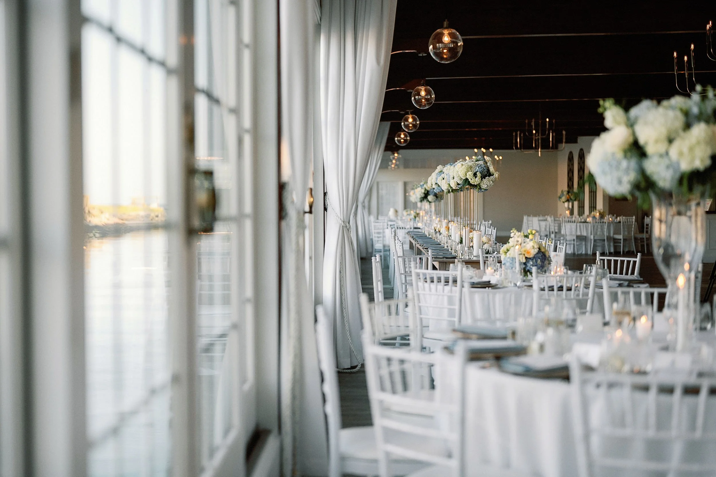 Elegant wedding reception setup with long tables, white chairs, and floral centerpieces in a bright room with large windows and draped curtains.