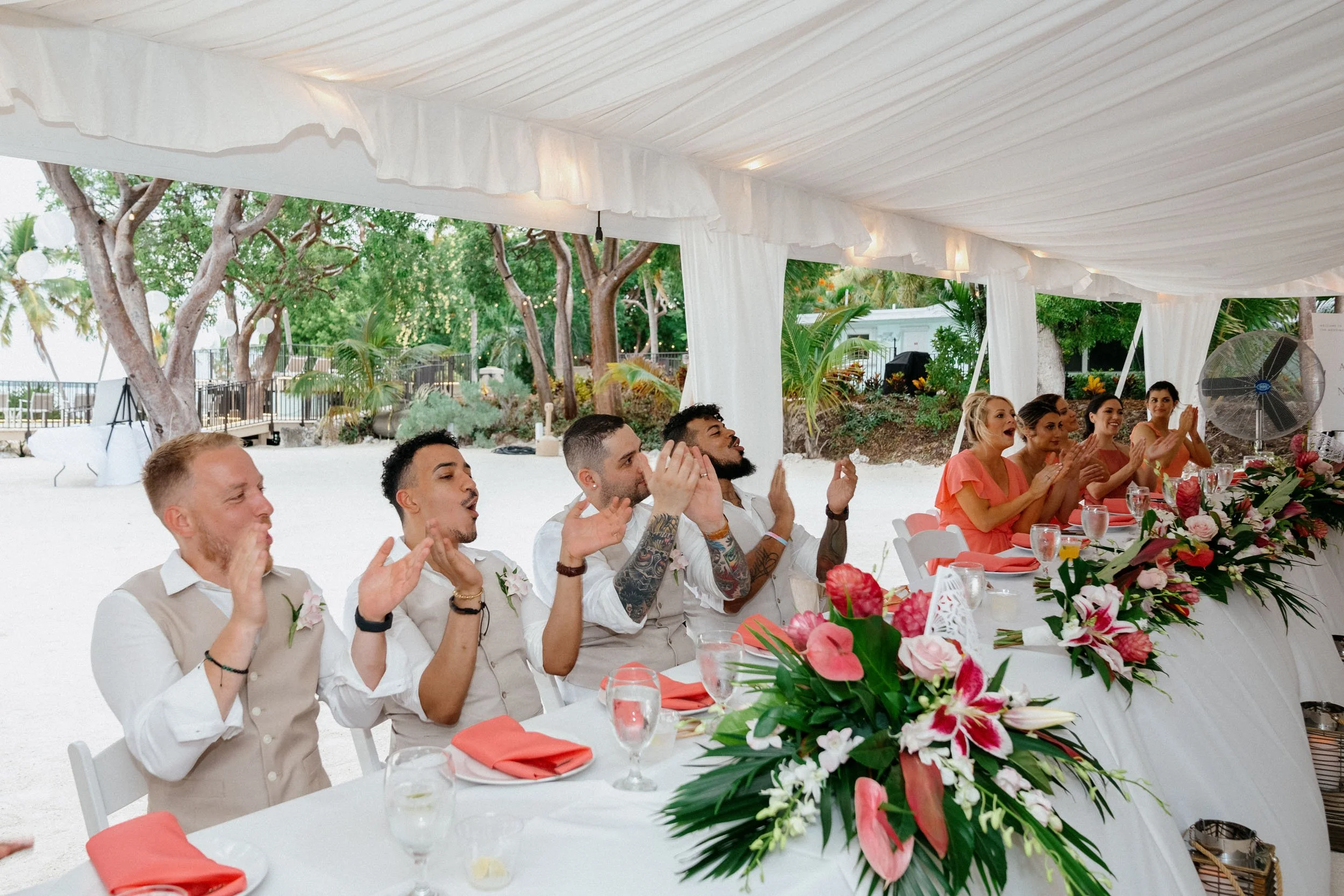 People sitting at a long decorated table inside a white tent with windows, clapping and enjoying a celebration, adorned with floral arrangements, with greenery and trees outside.