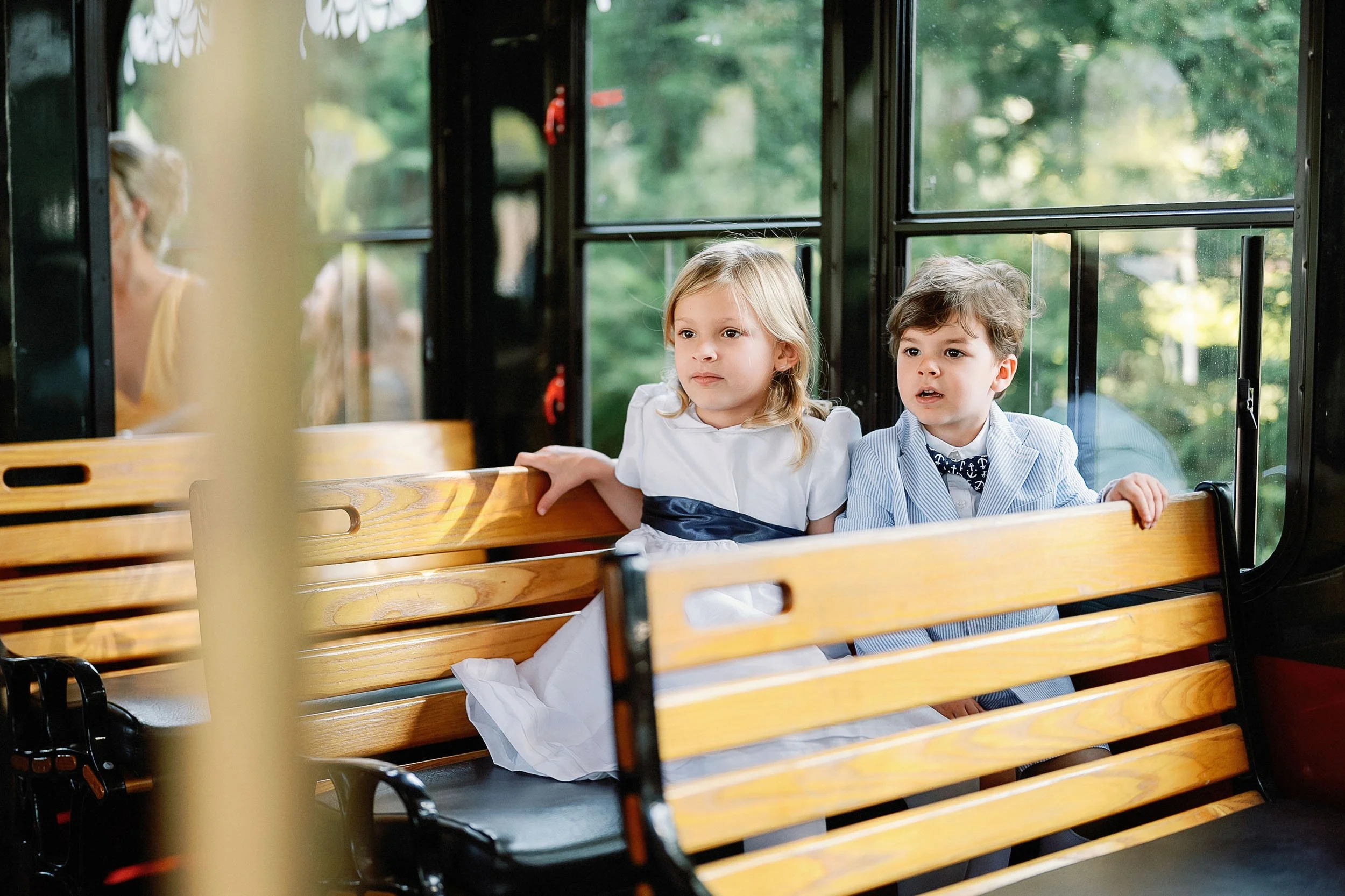 Two young children, a girl and a boy, sitting on a wooden bench inside a bus, looking out the window.