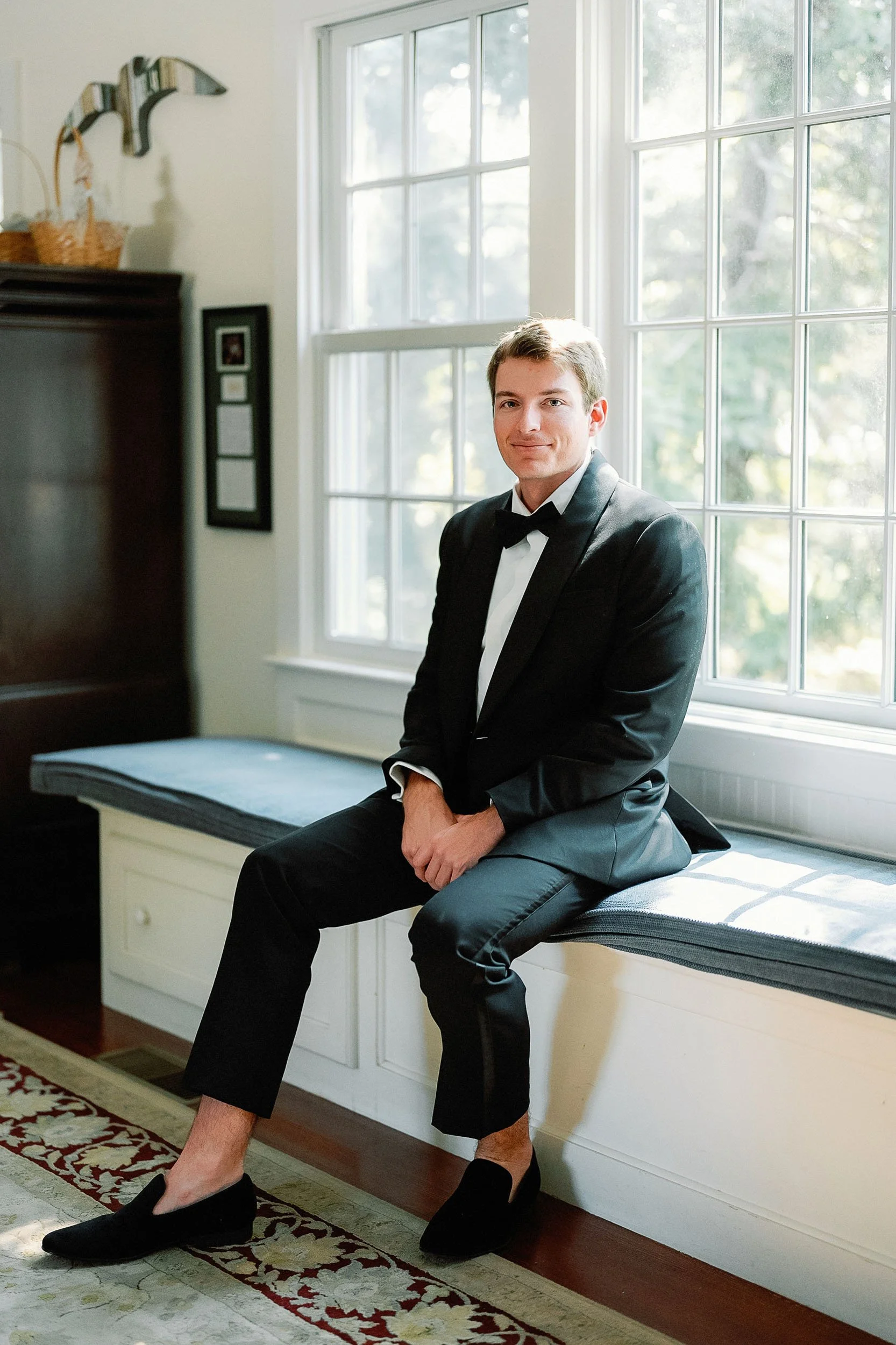 A young man in a tuxedo with a bow tie, sitting on a windowsill in a bright room, with sunlight coming through large multi-pane windows.