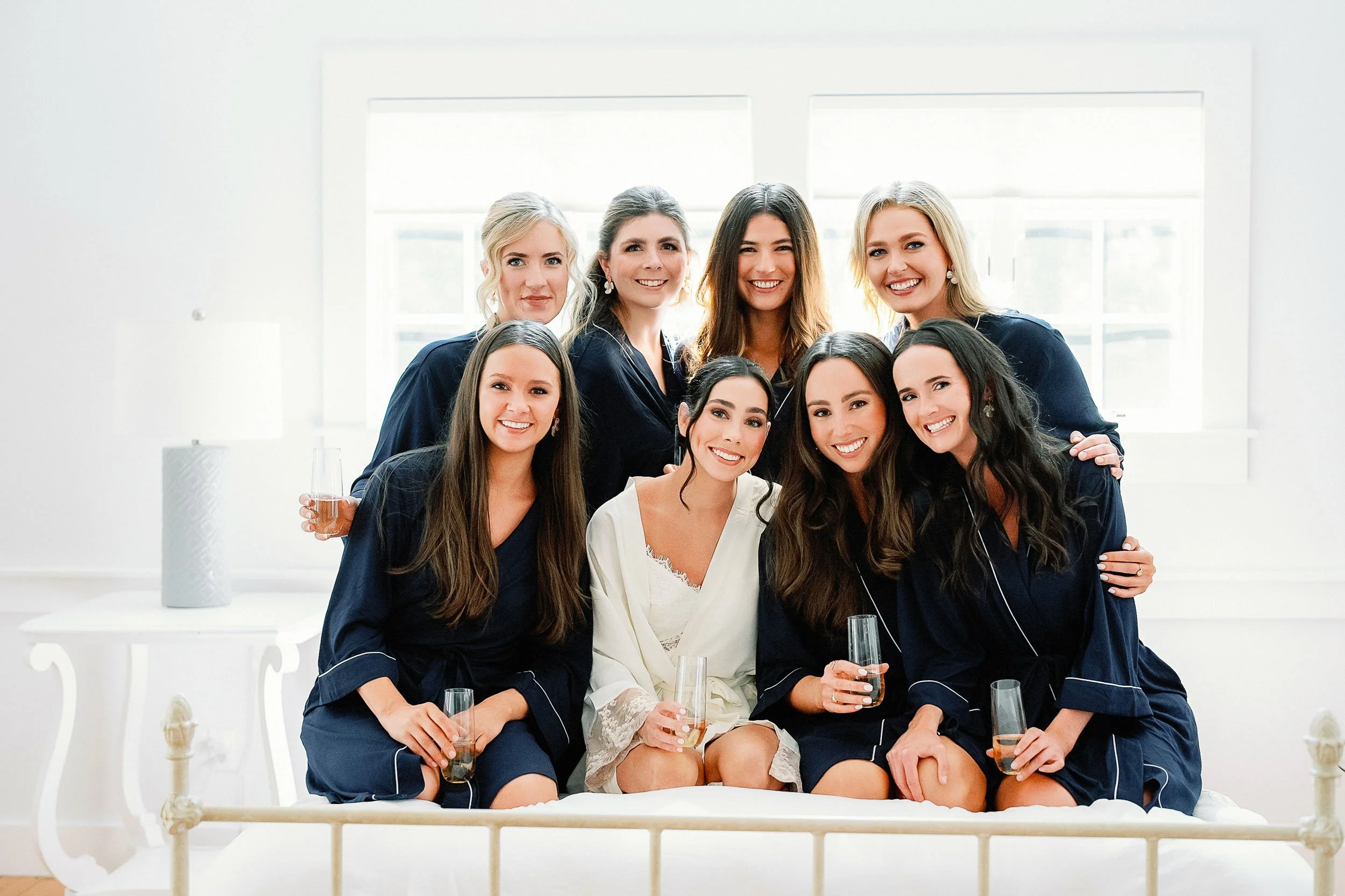 A group of nine women in pajamas celebrating, sitting on a bed with glasses of champagne, in a bright room with white walls and large windows.