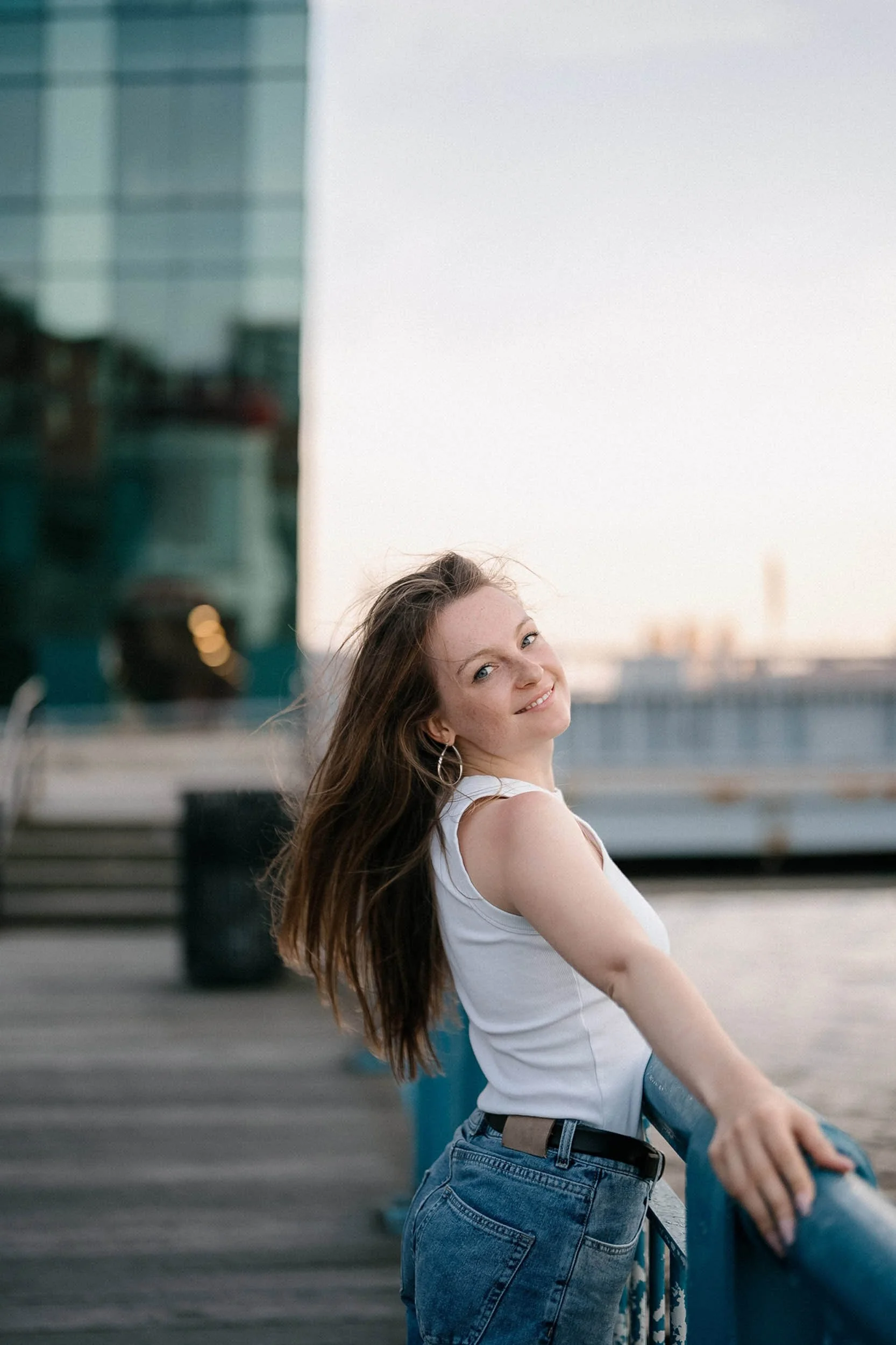 A young woman with long brown hair standing outdoors near water, smiling at the camera.