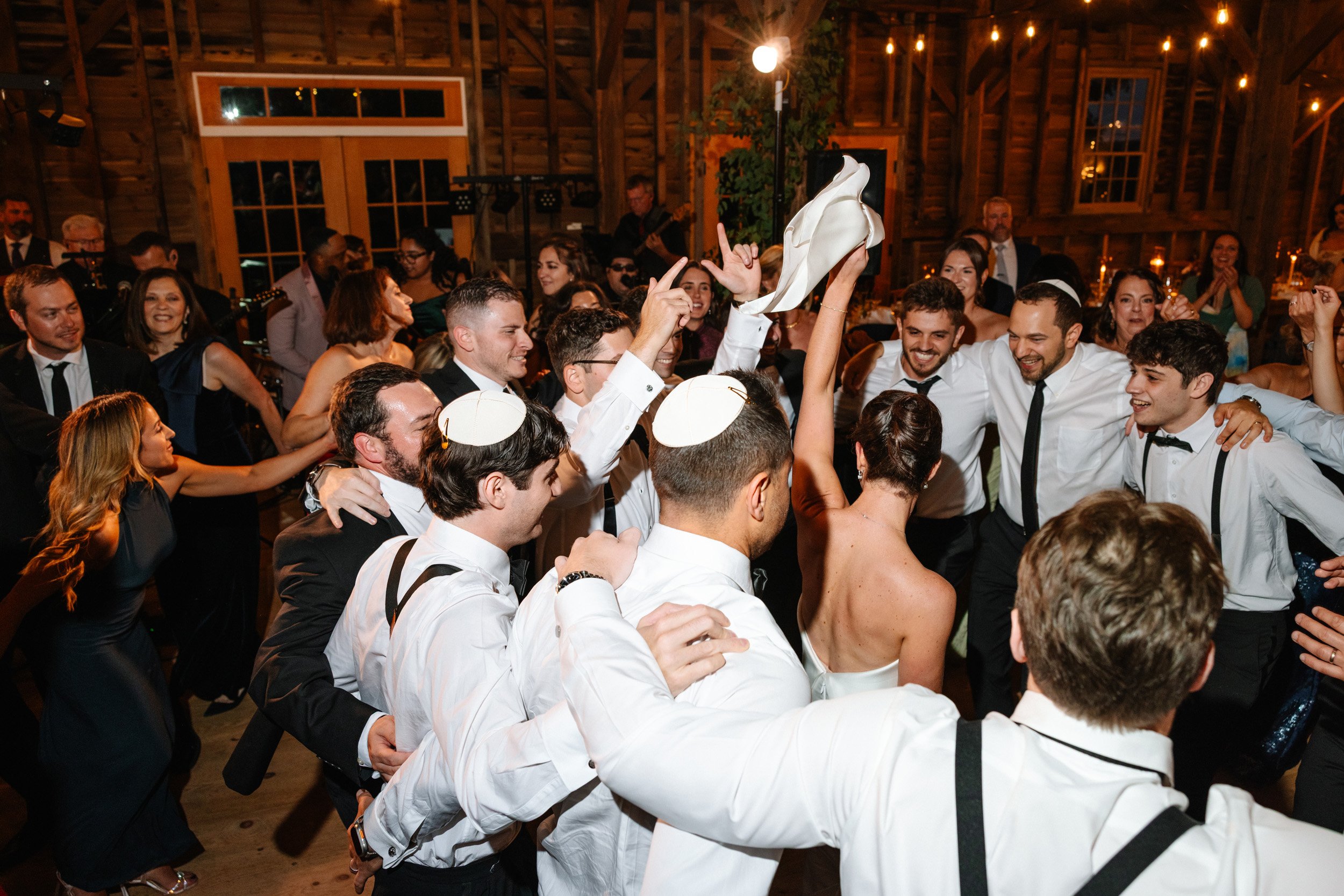 A wedding celebration with the bride and groom dancing and surrounded by friends and family, with some wearing kippahs, in a rustic wooden venue with string lights.